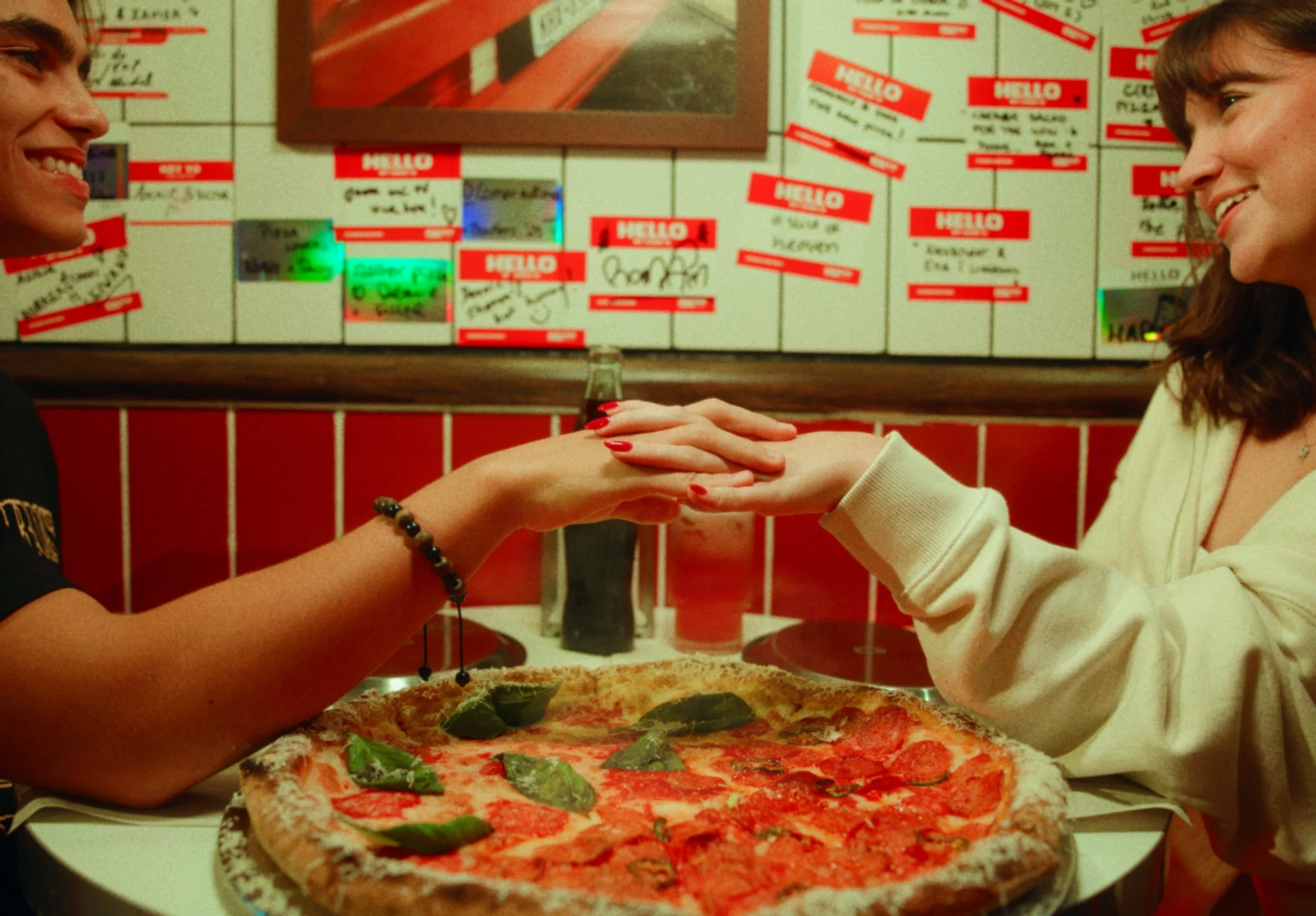 A couple sitting at a table with a pizza, holding hands and smiling at each other in a restaurant with red and white decor and a wall covered with red
