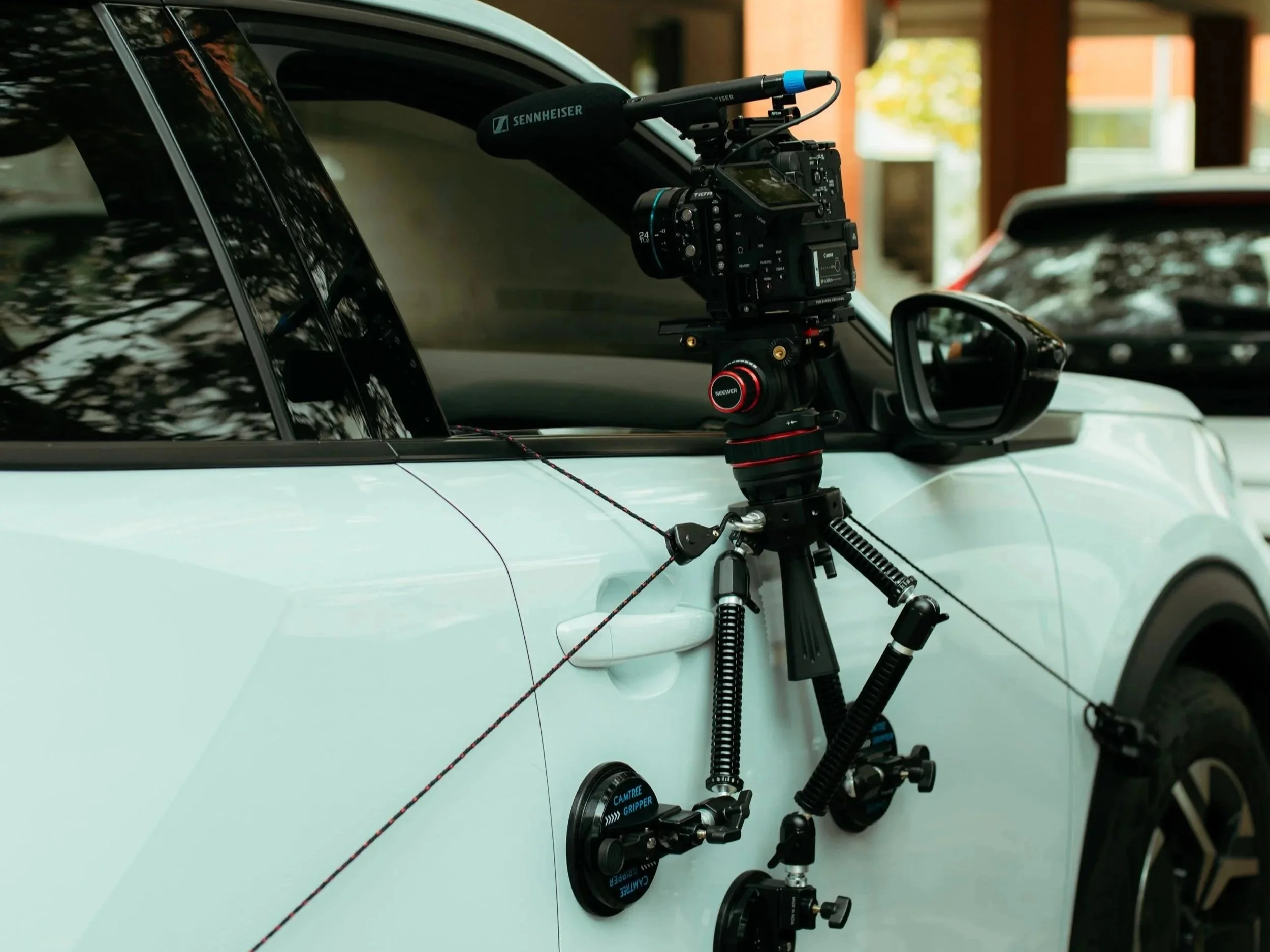 Camera mounted on a tripod attached to the side of a white vehicle with a suction cup. The camera has a microphone labeled 'Sennheiser' and is positioned next to the vehicle window.