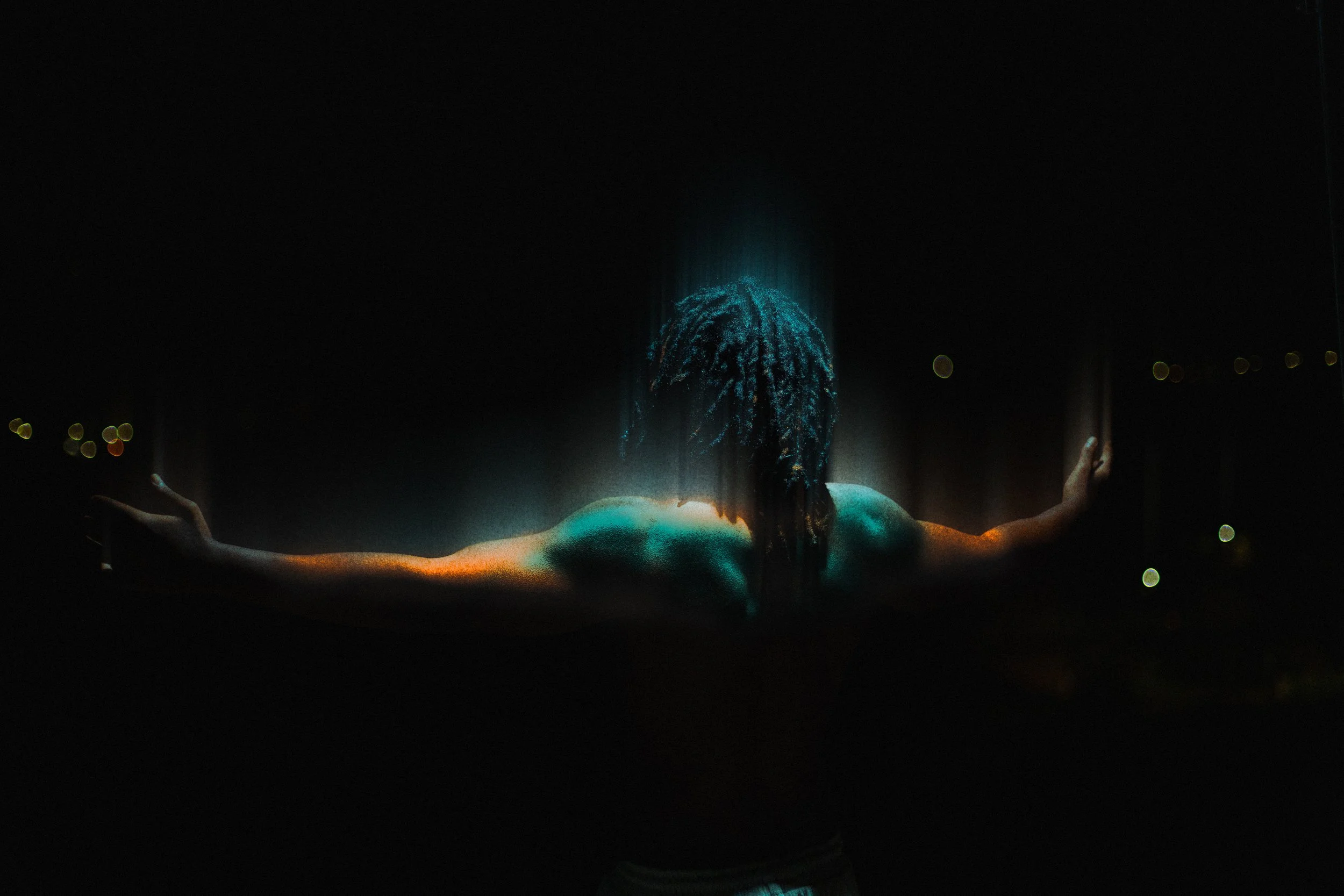 Person with dreadlocks facing away, arms outstretched, illuminated in a dark environment with city lights in the background.