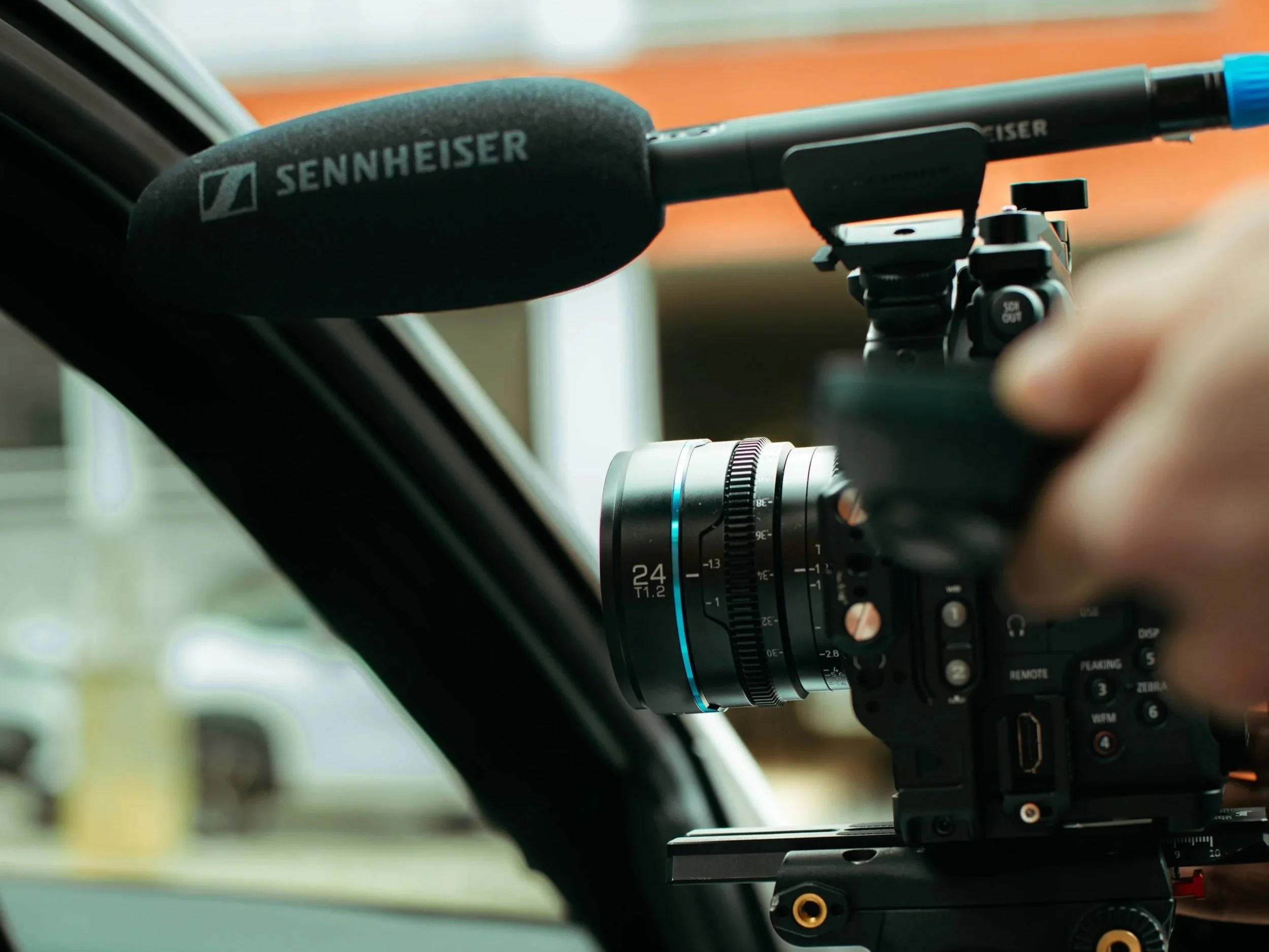 Close-up of a videographer adjusting a professional camera with a focus lens, inside a vehicle, with a Sennheiser microphone attached.