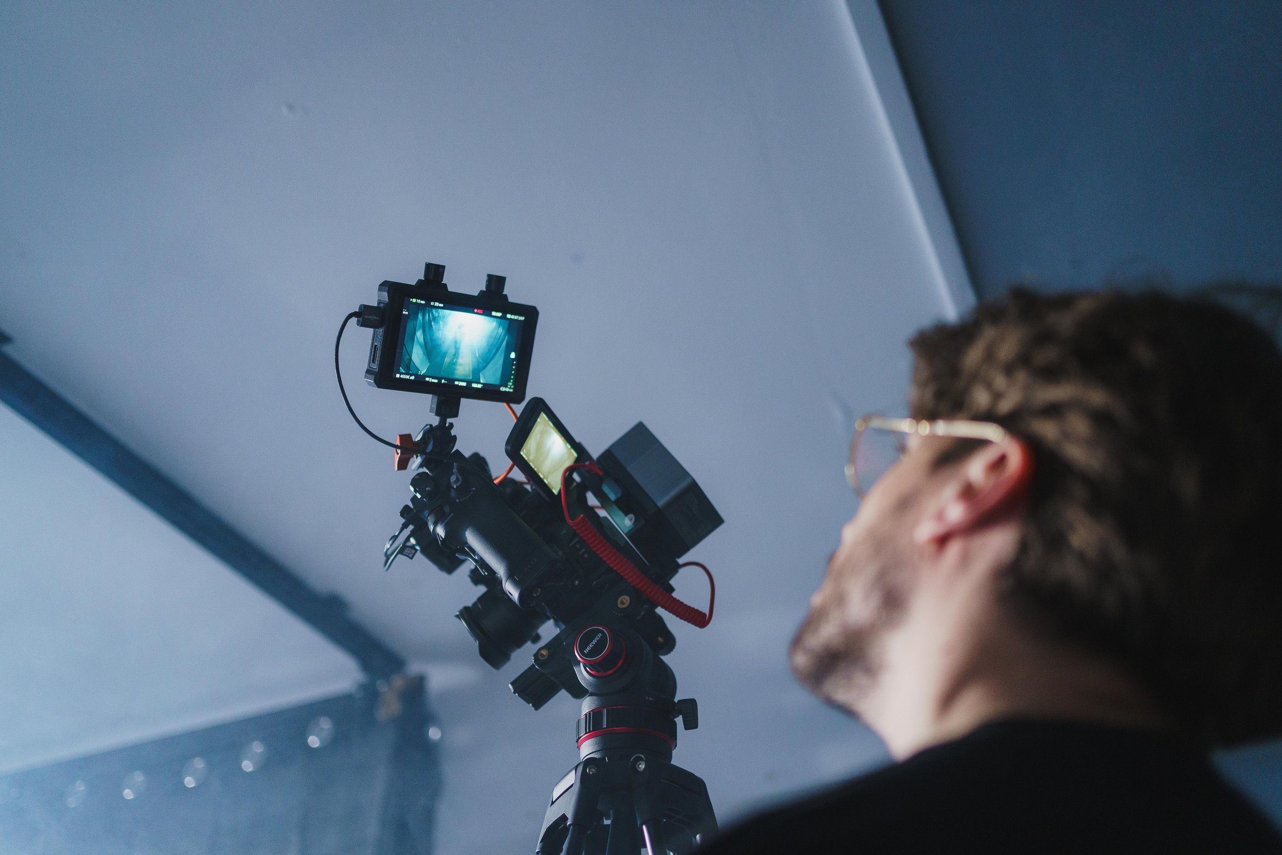 A person with glasses operating a professional camera on a tripod, capturing footage or images indoors.