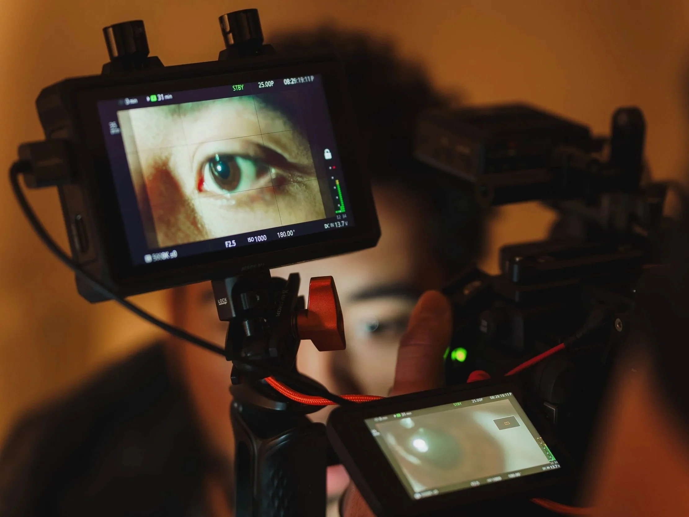 Close-up of a camera monitor displaying a detailed view of a human eye with long eyelashes and light-colored skin, with a person operating the camera in the background.