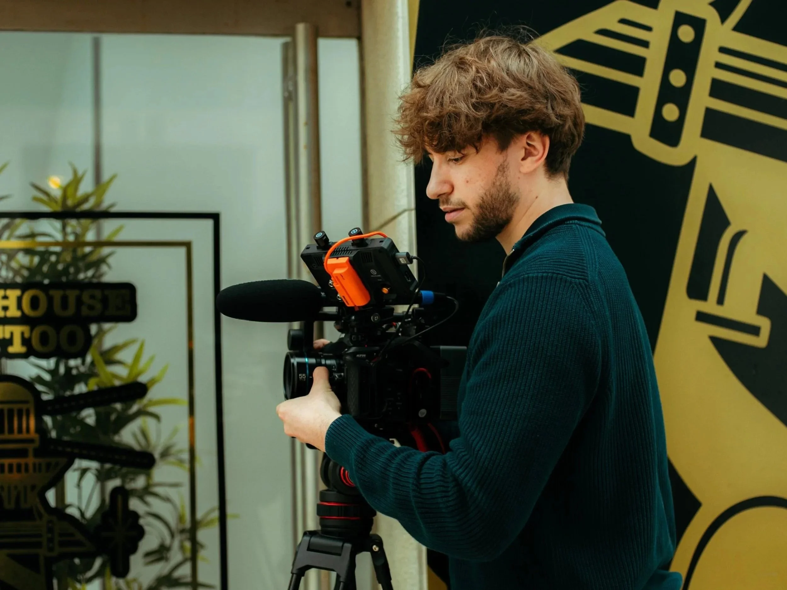 Young man with curly hair and beard operating professional video camera on tripod at an indoor event.