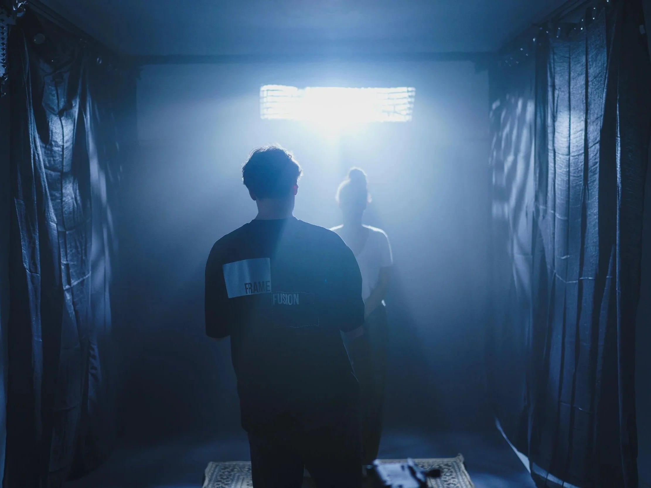 People inside a dark room with blue lighting and curtains on the sides, with a bright light source behind them creating silhouettes.