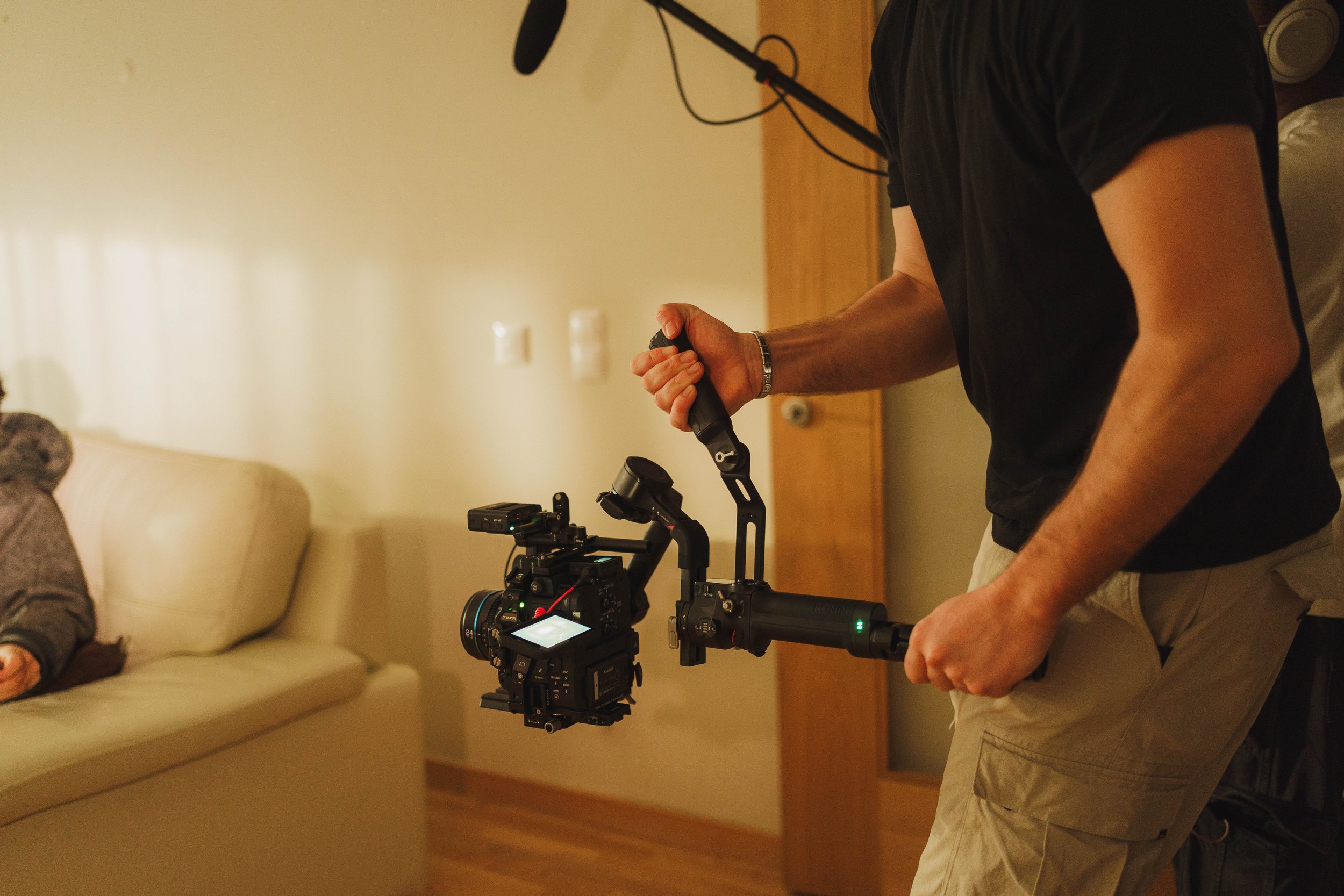 Person operating a professional camera stabilizer mounted on a camera inside a room, with a partial view of a sofa and another person sitting on it.