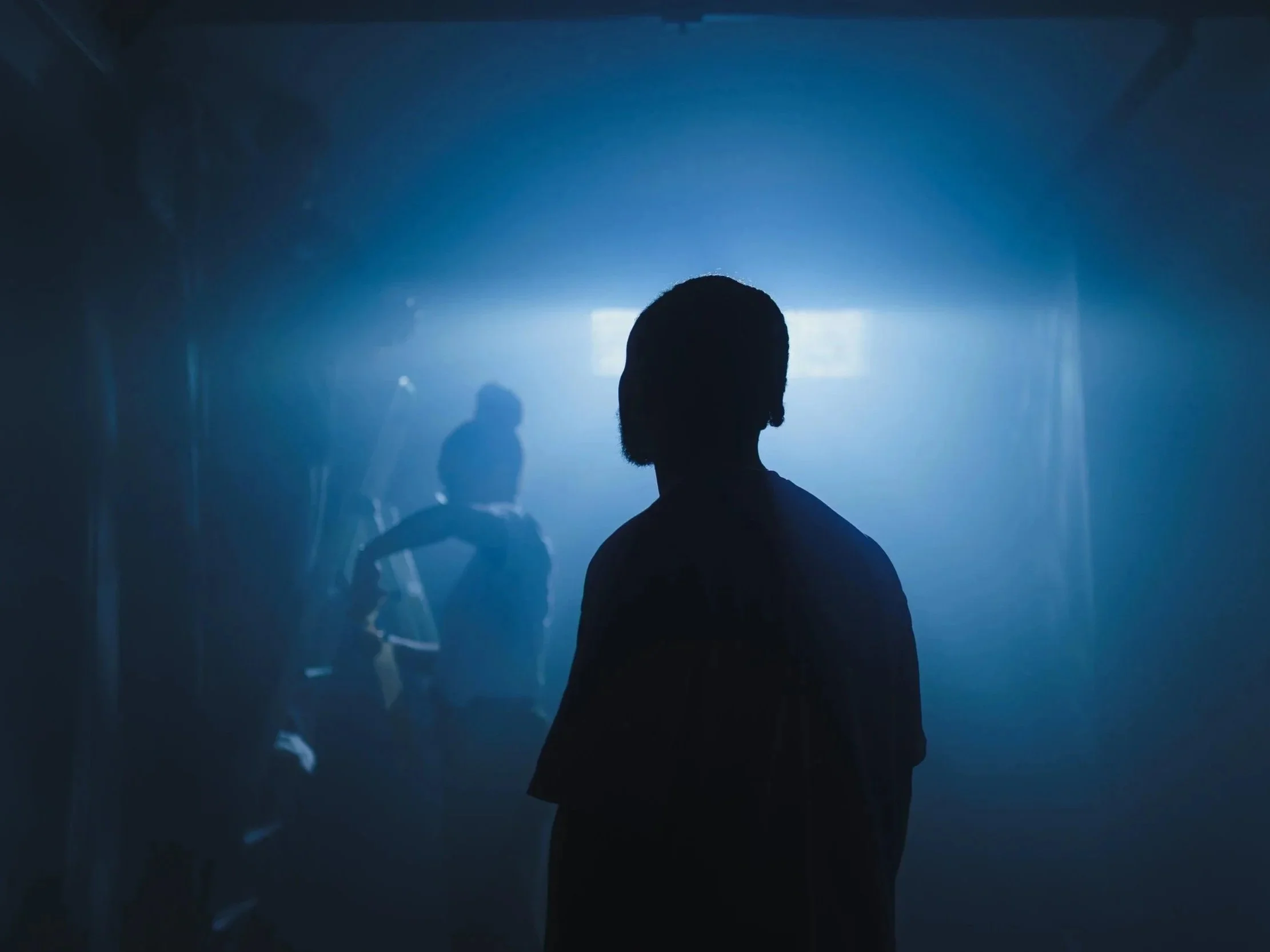 Silhouette of a person standing in front of a foggy, dimly lit room with two figures sitting on ladders in the background