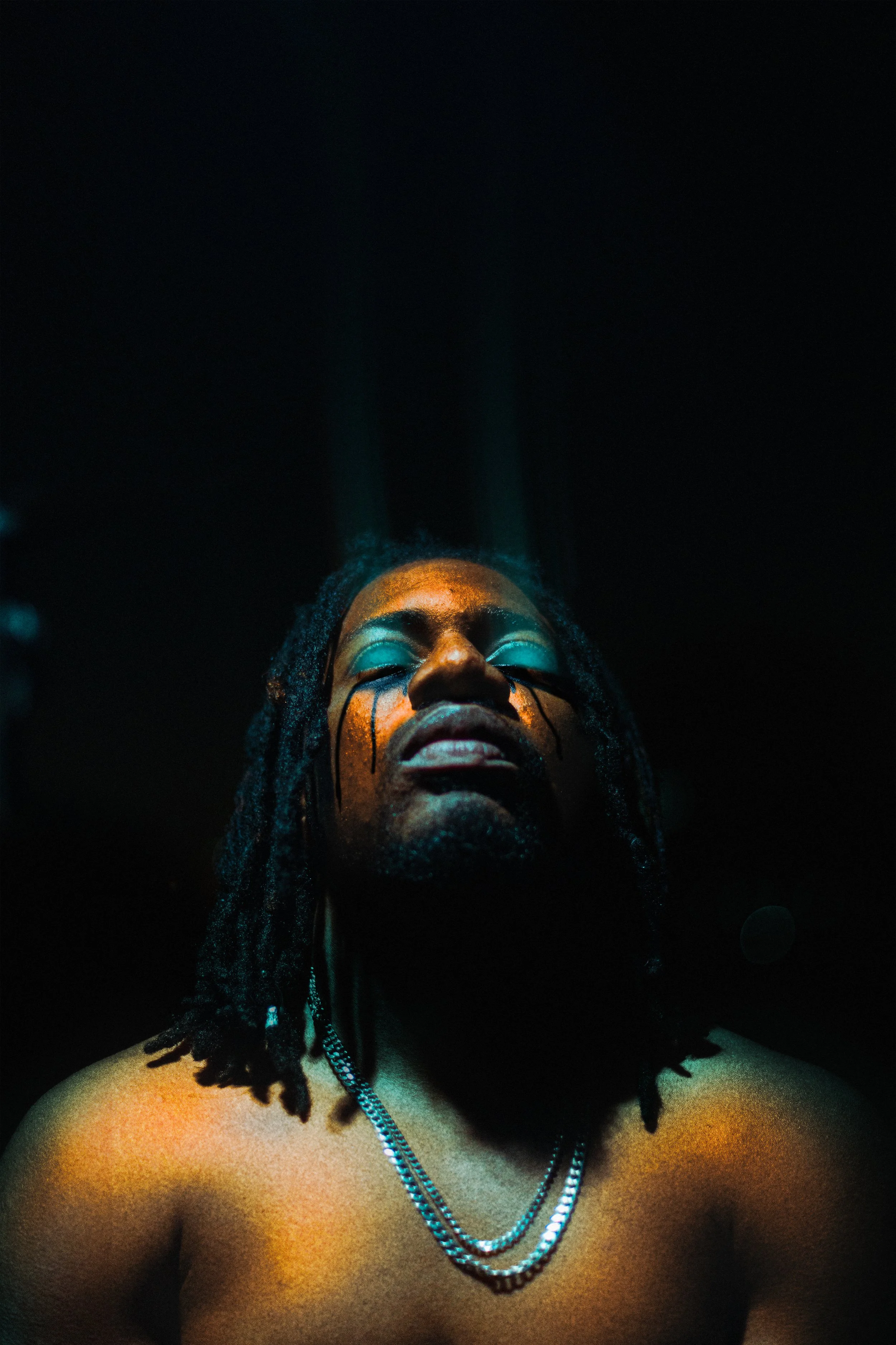 A shirtless man with long dreadlocks and a silver chain, with a painted face expressing intense emotion, illuminated by dramatic blue and orange lighting in a dark setting.