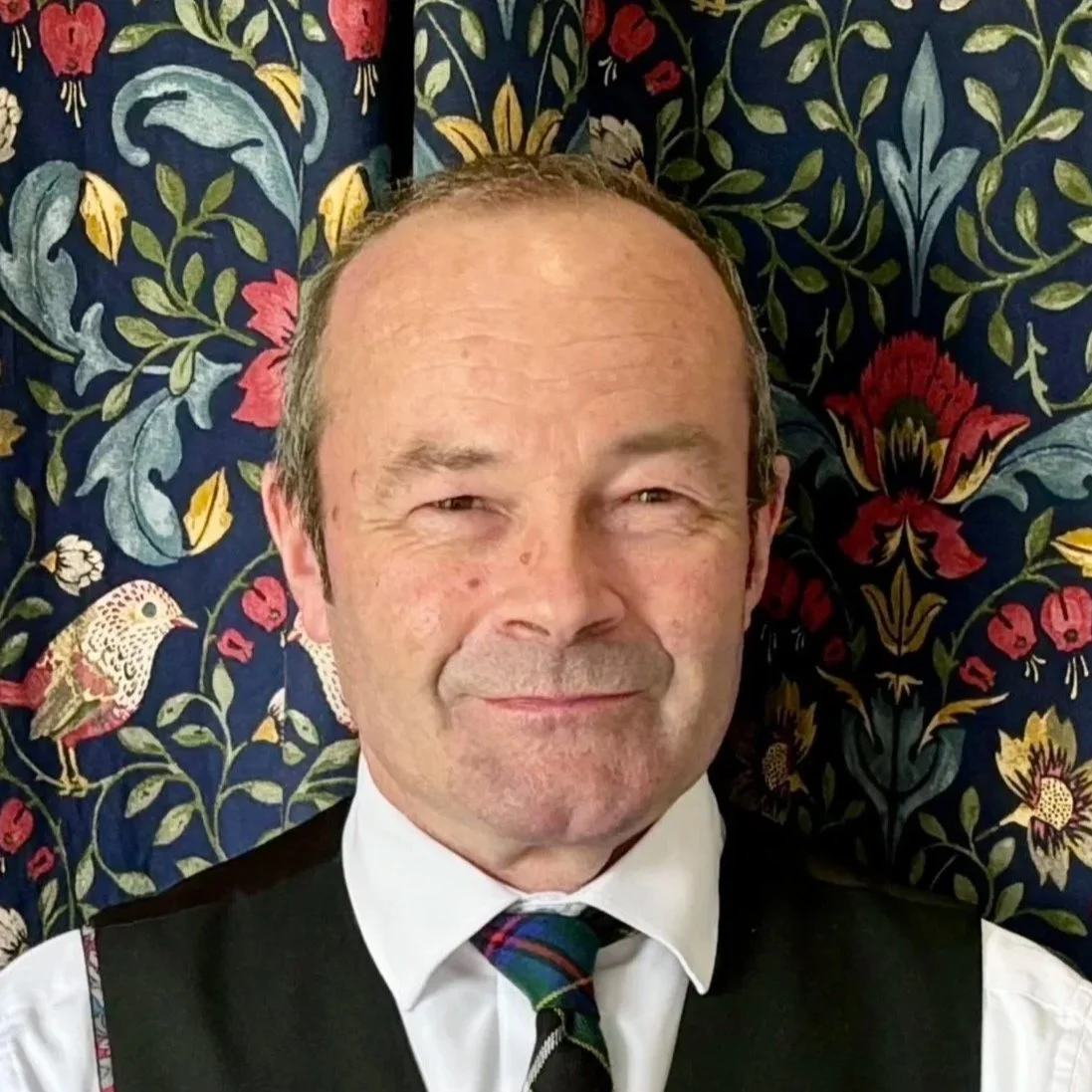 A smiling middle-aged man with short hair, wearing a white shirt, a black vest, and a colorful striped tie, standing in front of a navy blue fabric with a floral pattern and birds.