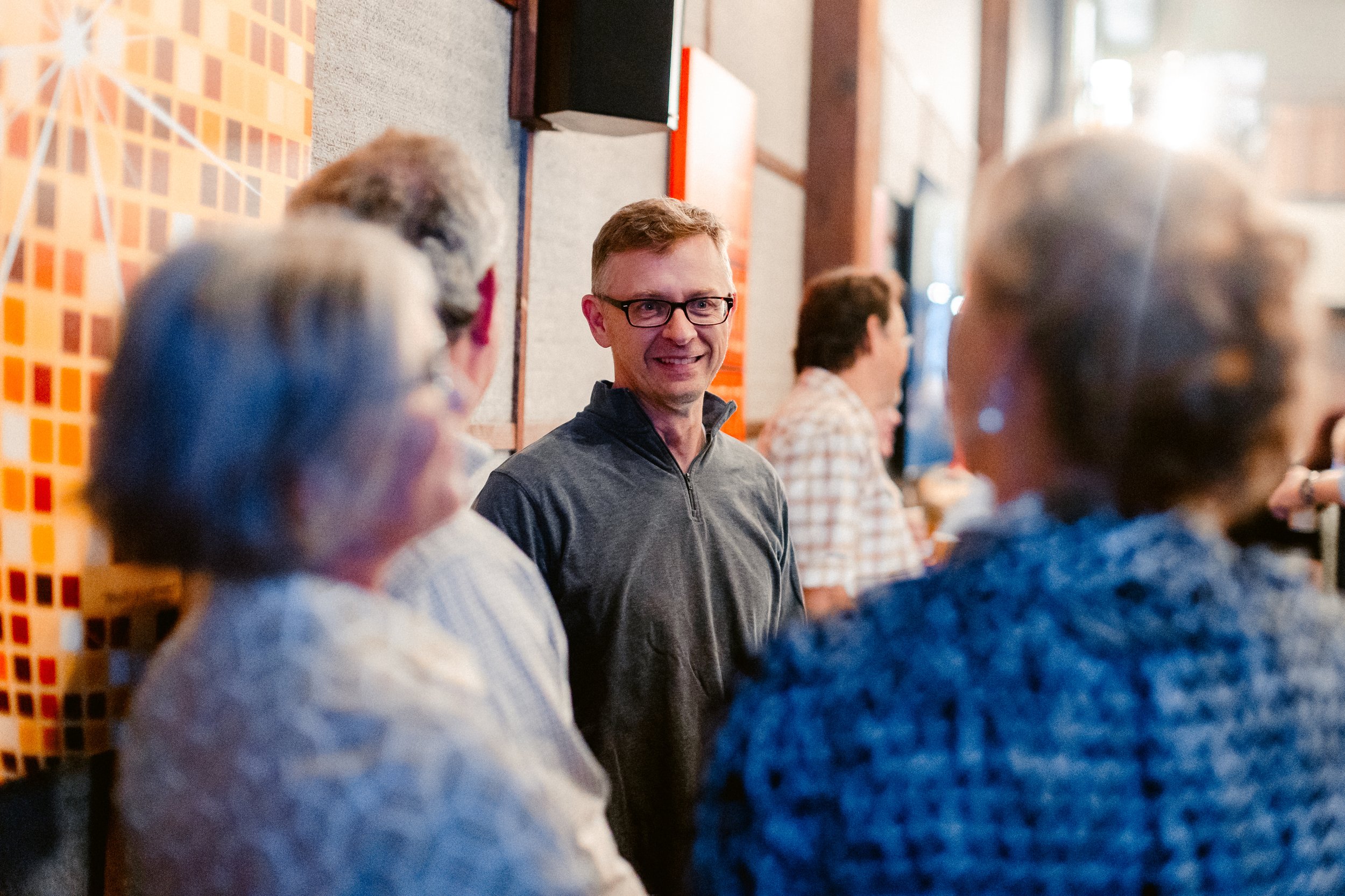 Man smiling and talking to a group of women in a social setting, with other people in the background.