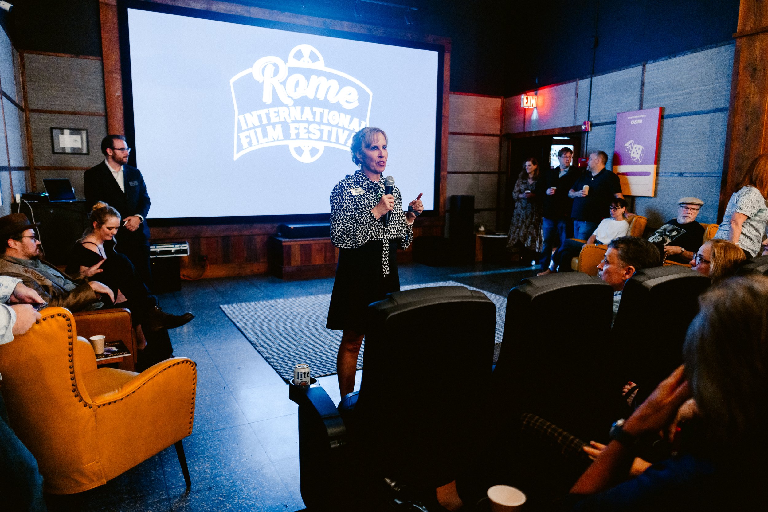 A woman is speaking into a microphone in a room with a large screen that reads 'Rome International Film Festival'. Several people are seated and standing, listening to her, with some taking photos or using their phones.