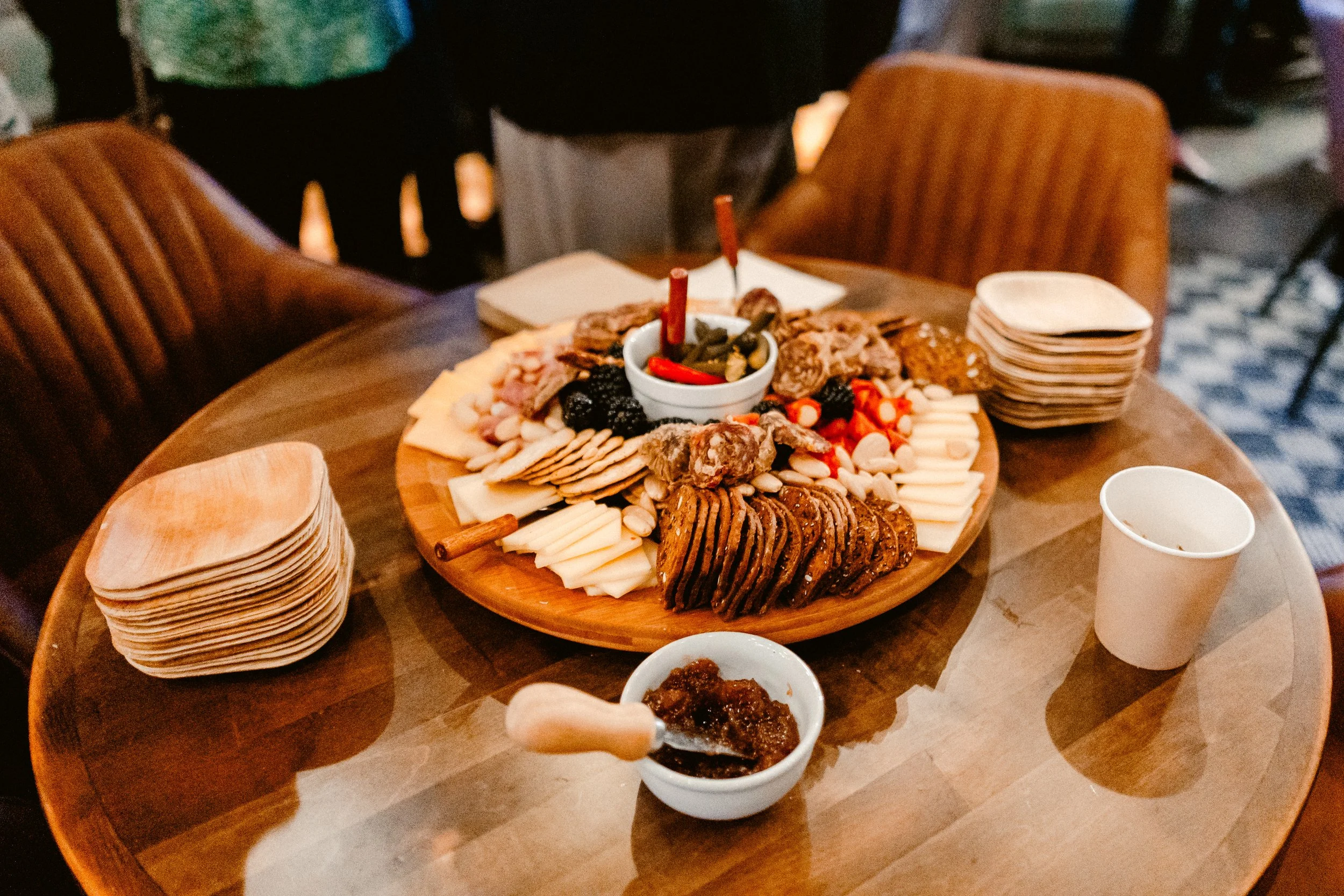 A round wooden table with a charcuterie board containing assorted cheeses, meats, crackers, and fruit, along with a small bowl of chutney or jam, paper napkins, and disposable cups in a cozy restaurant setting.