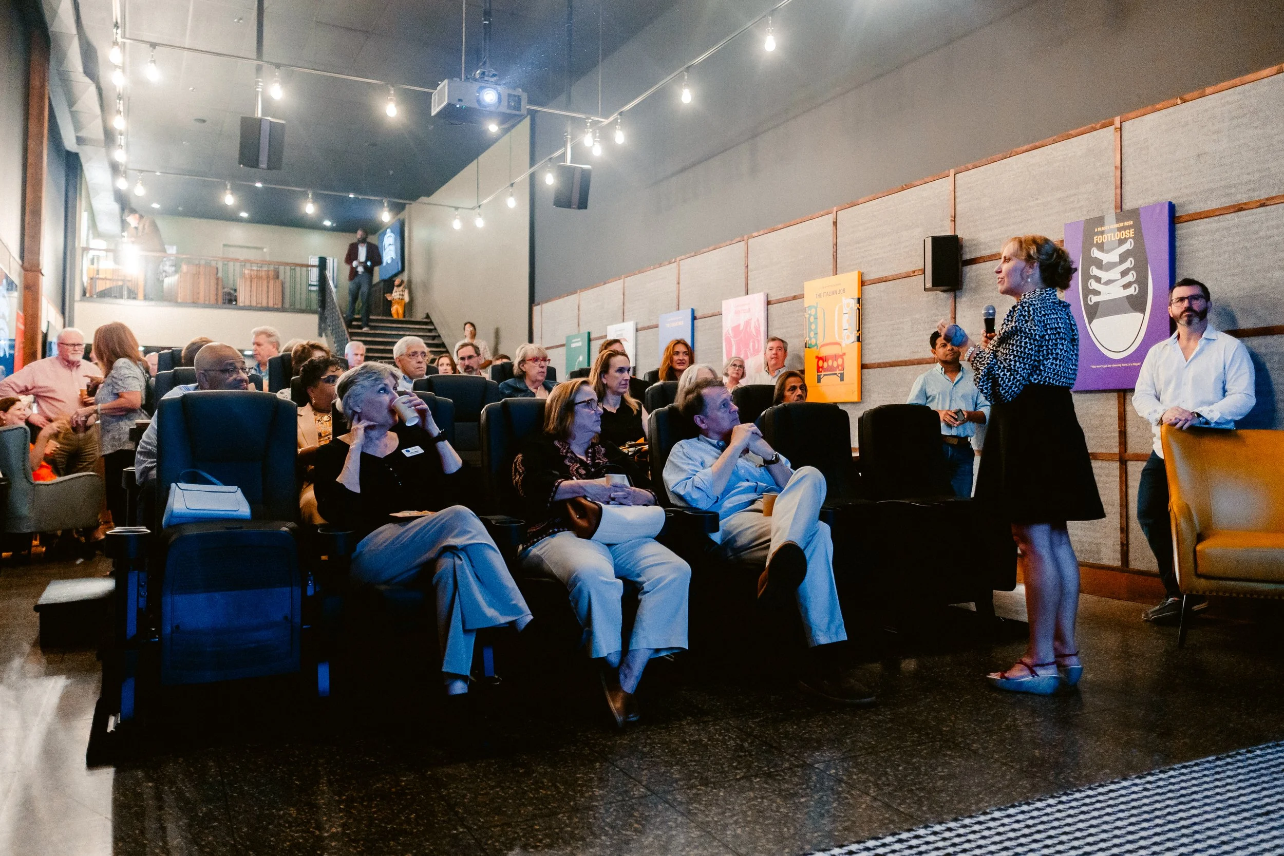 A woman holding a microphone and speaking to an audience seated in a modern conference room with artwork on the wall.