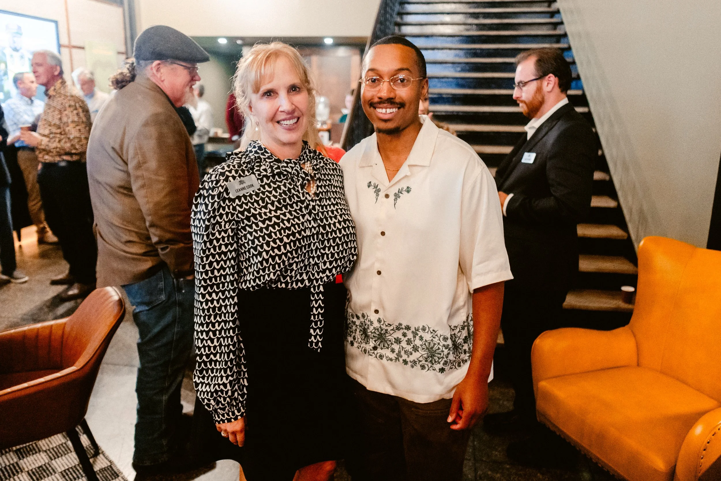 A woman and a man smiling at a social event, standing in front of seating with other people mingling in the background.