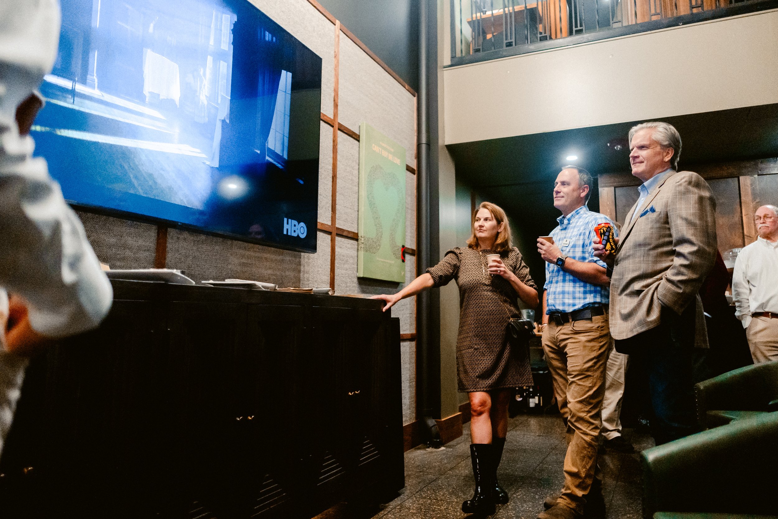 Group of four people watching a television, with one person pointing at the screen, in a casual indoor setting.