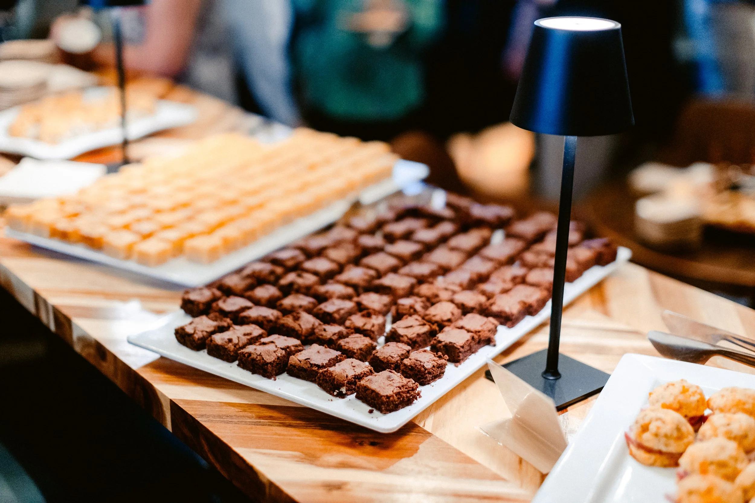 Tray of chocolate brownies arranged on a white rectangular platter at a dessert buffet, with other desserts blurred in the background.