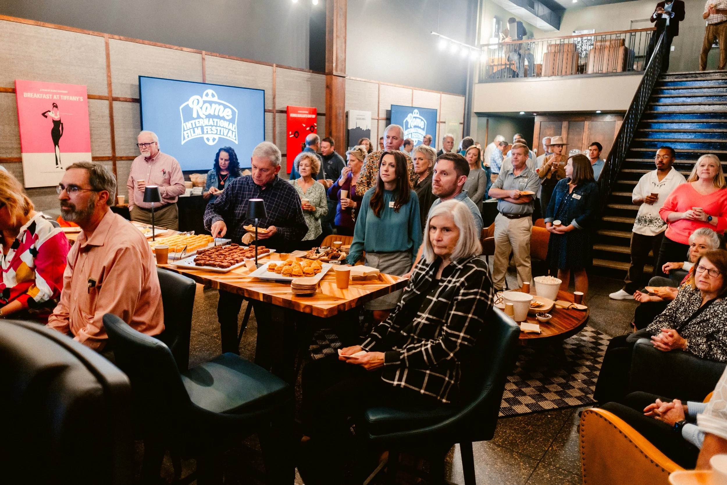 People gathered in a room for a film festival event with food and drinks, with screens displaying 'Rome International Film Festival' in the background.