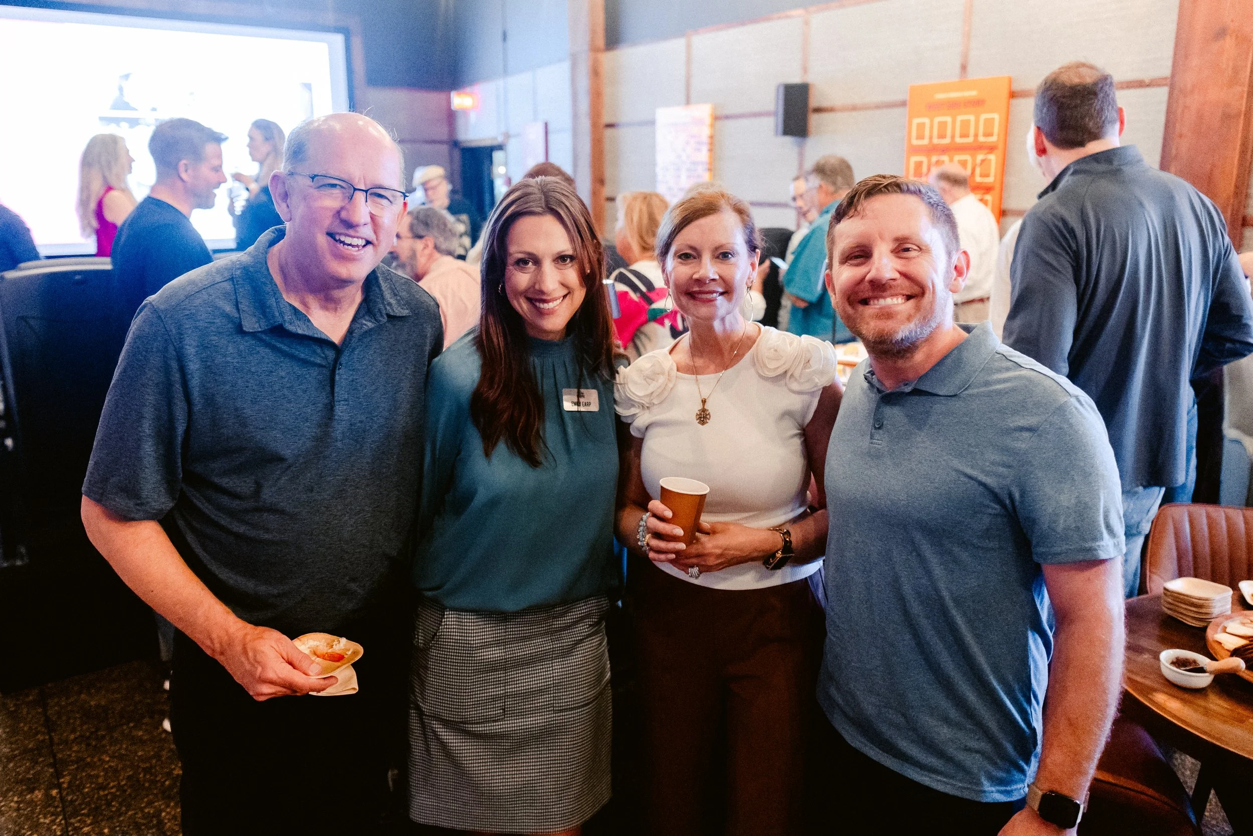 Four adults smiling and standing together in a crowded indoor event space, with people in the background, some holding drinks, and a large screen on the wall.