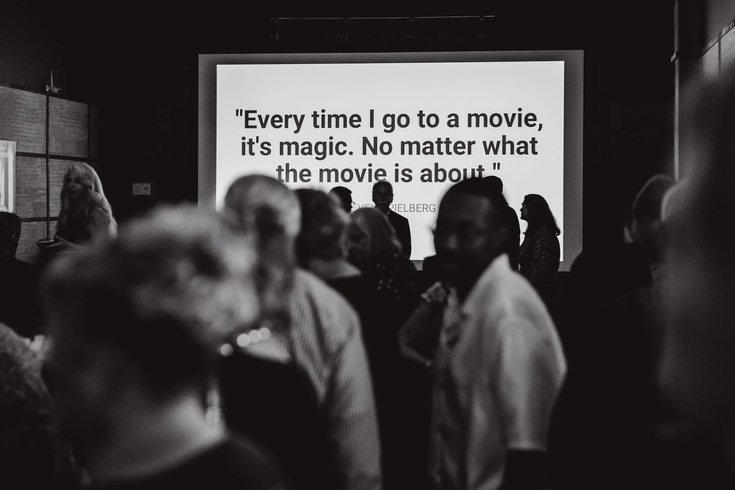 People attending a movie screening or presentation, with a large screen displaying a quote by Steven Spielberg, in black and white.