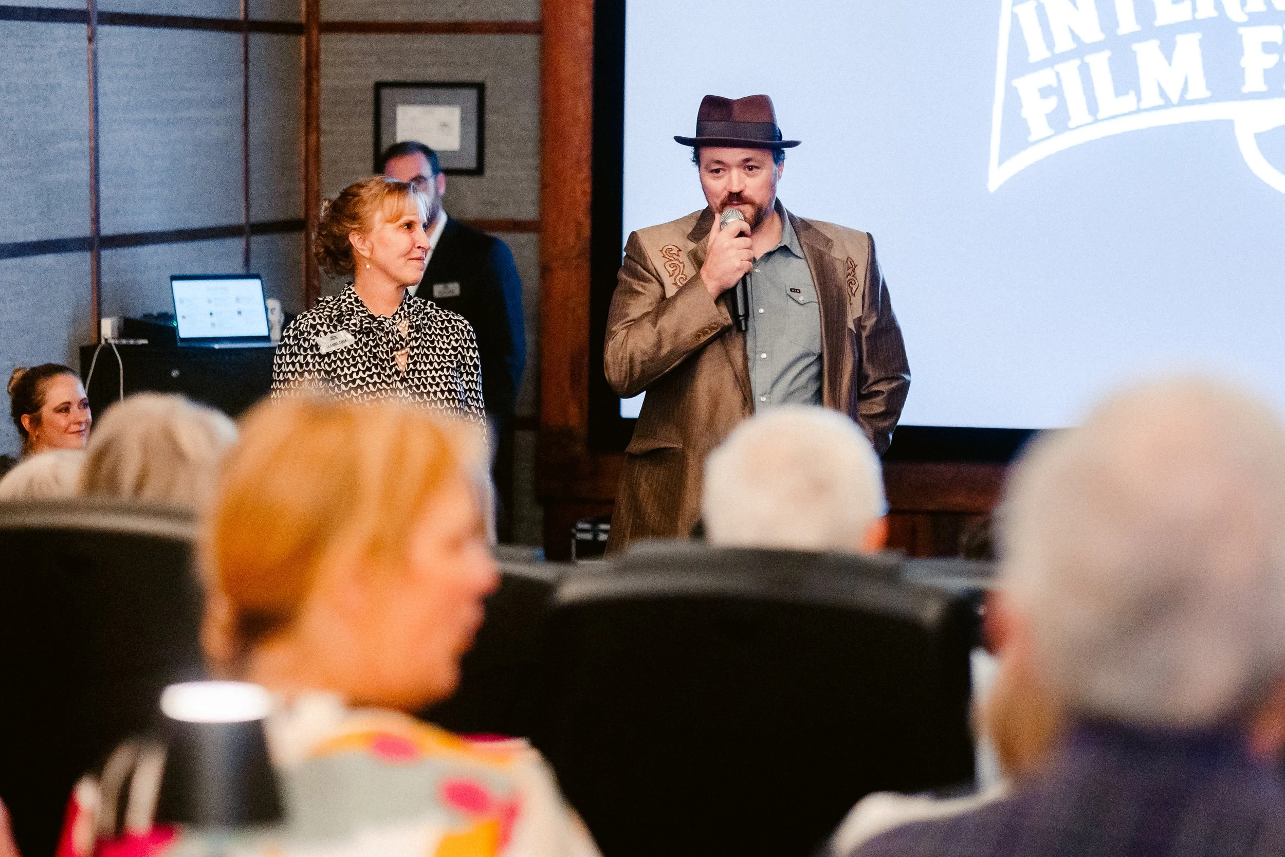 A man in a brown jacket and fedora speaking into a microphone at a conference or event, standing in front of a large screen displaying the words 'International Film.'