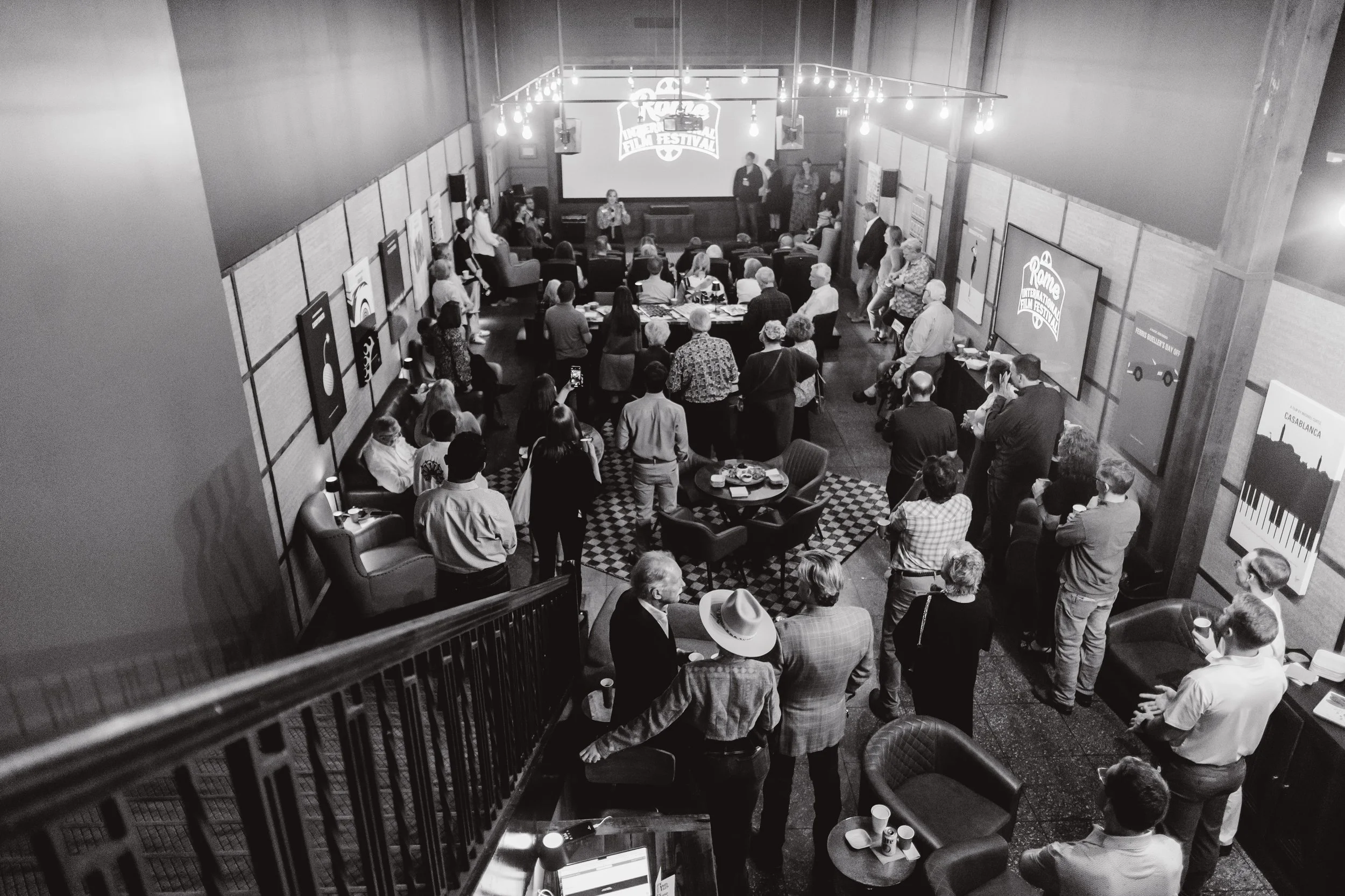 People attending a presentation in a room with large screens displaying the logo 'Rome Film Festival'. The audience is seated and standing, watching the speaker on stage amidst framed artwork on the walls.