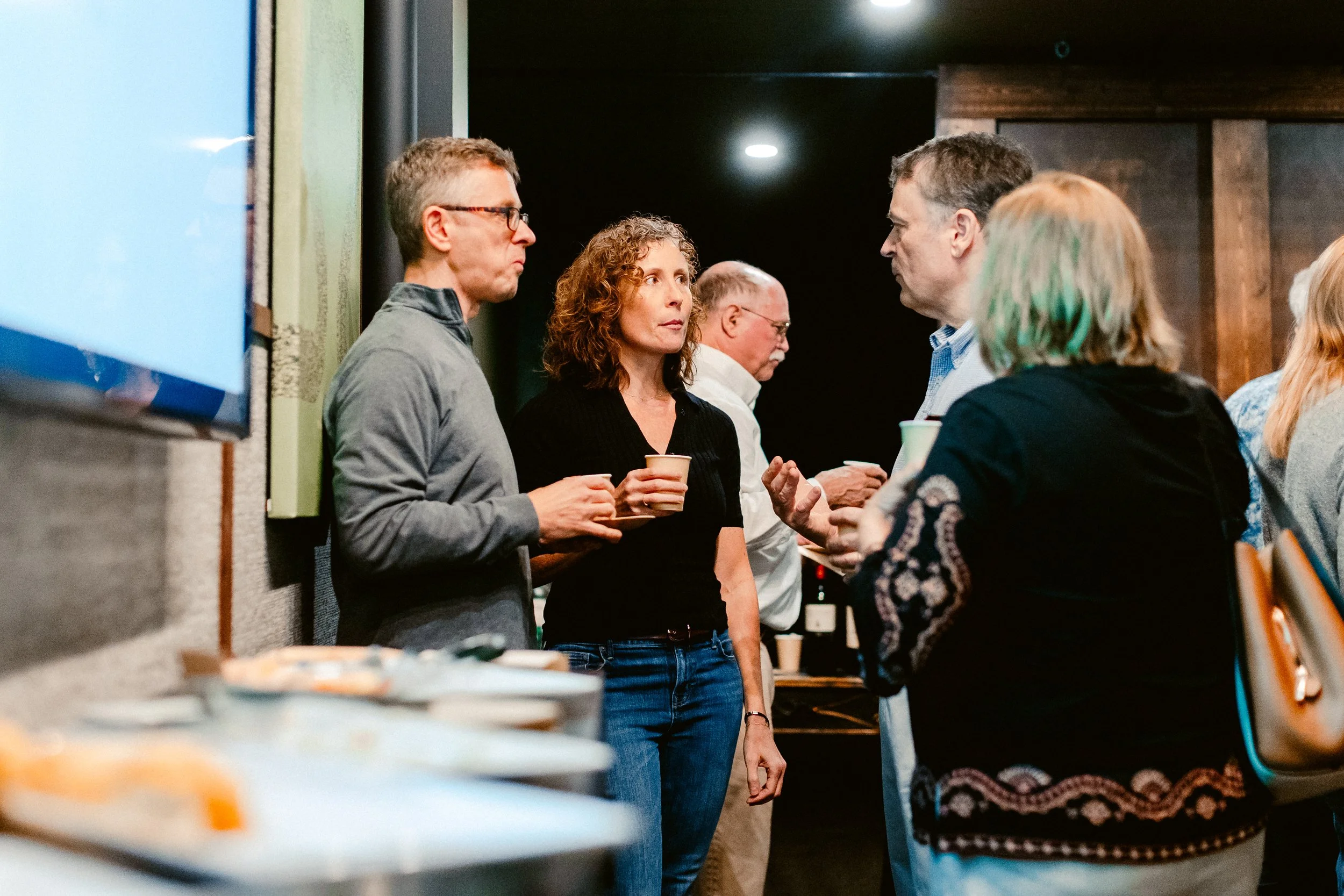 Group of people engaging in conversation at a social gathering, with some holding cups, in an indoor setting.