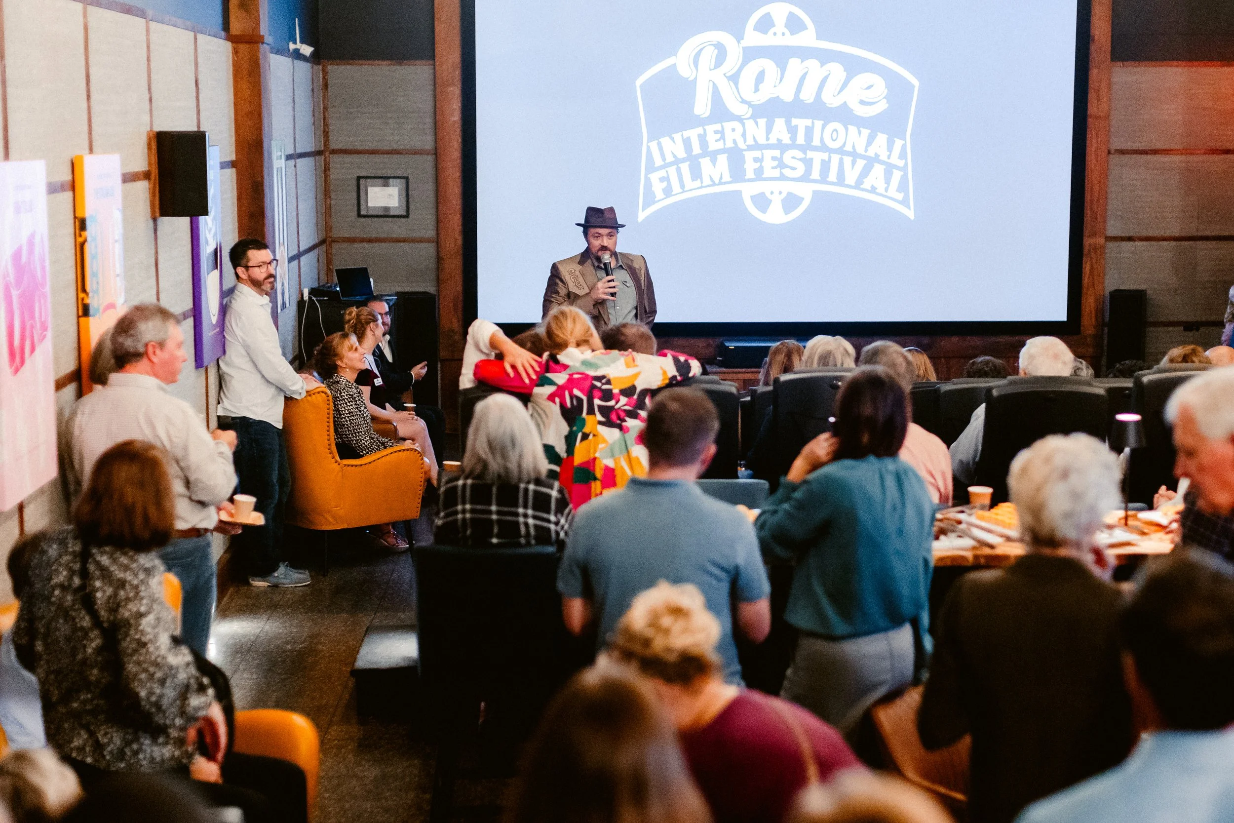 A man in a hat and blazer speaking into a microphone at the Rome International Film Festival, with an audience seated and standing around him. A large screen in the background displays the festival's logo.