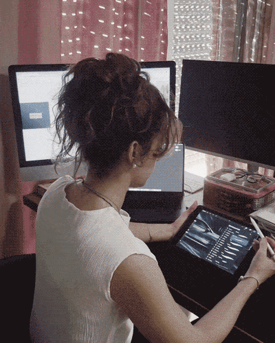 Una donna seduta a un tavolo con tre schermi e un tablet, lavora o studia, con luci soffuse e tende rosa sullo sfondo.