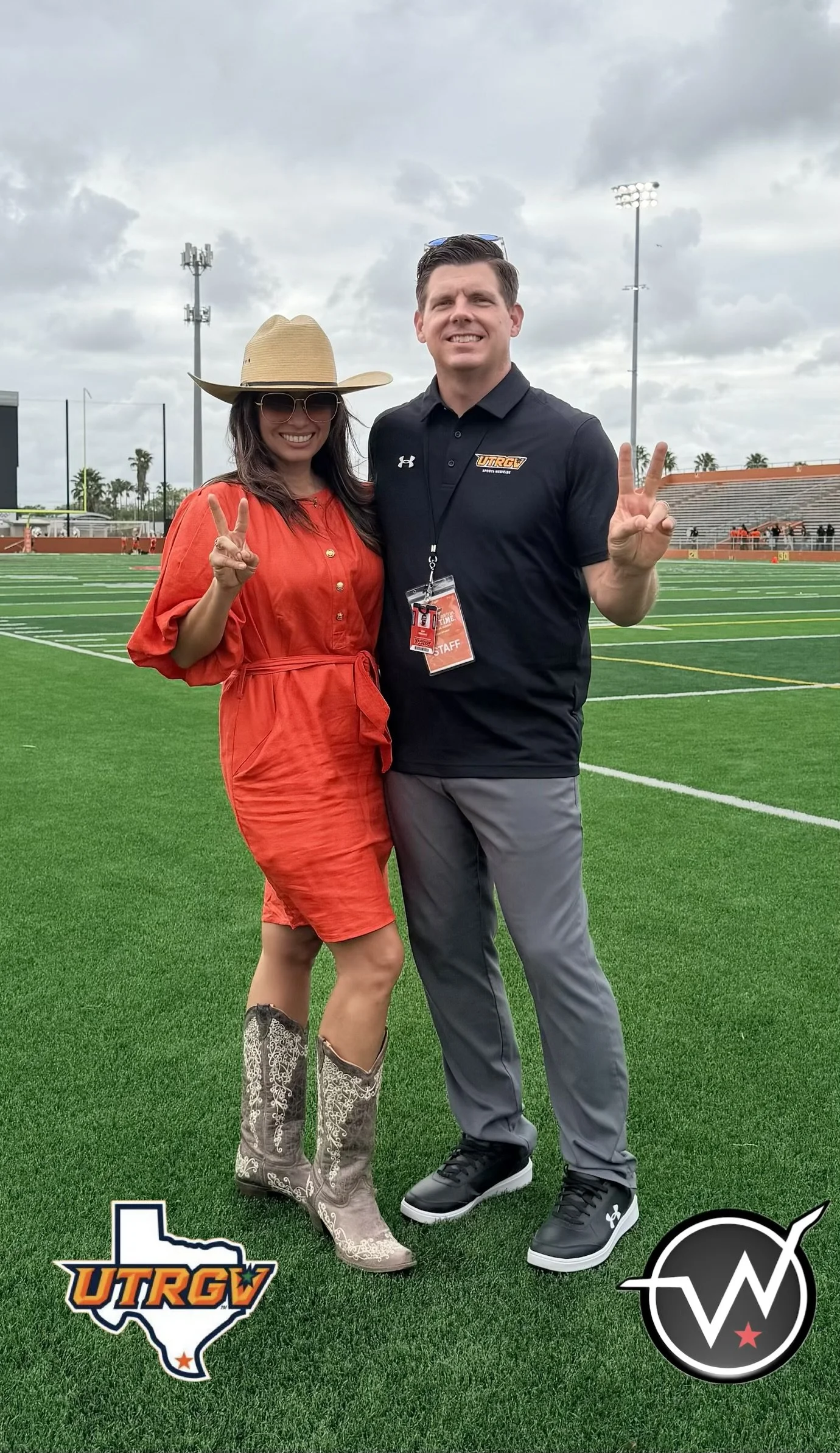 A man and woman standing on a football field, smiling and making peace signs. The woman is wearing a large cowboy hat, sunglasses, a red dress, and cowboy boots. The man is wearing a black polo shirt with the logo "UTRGV" and gray pants. There are football field markings, a cloudy sky, and stadium lights in the background.