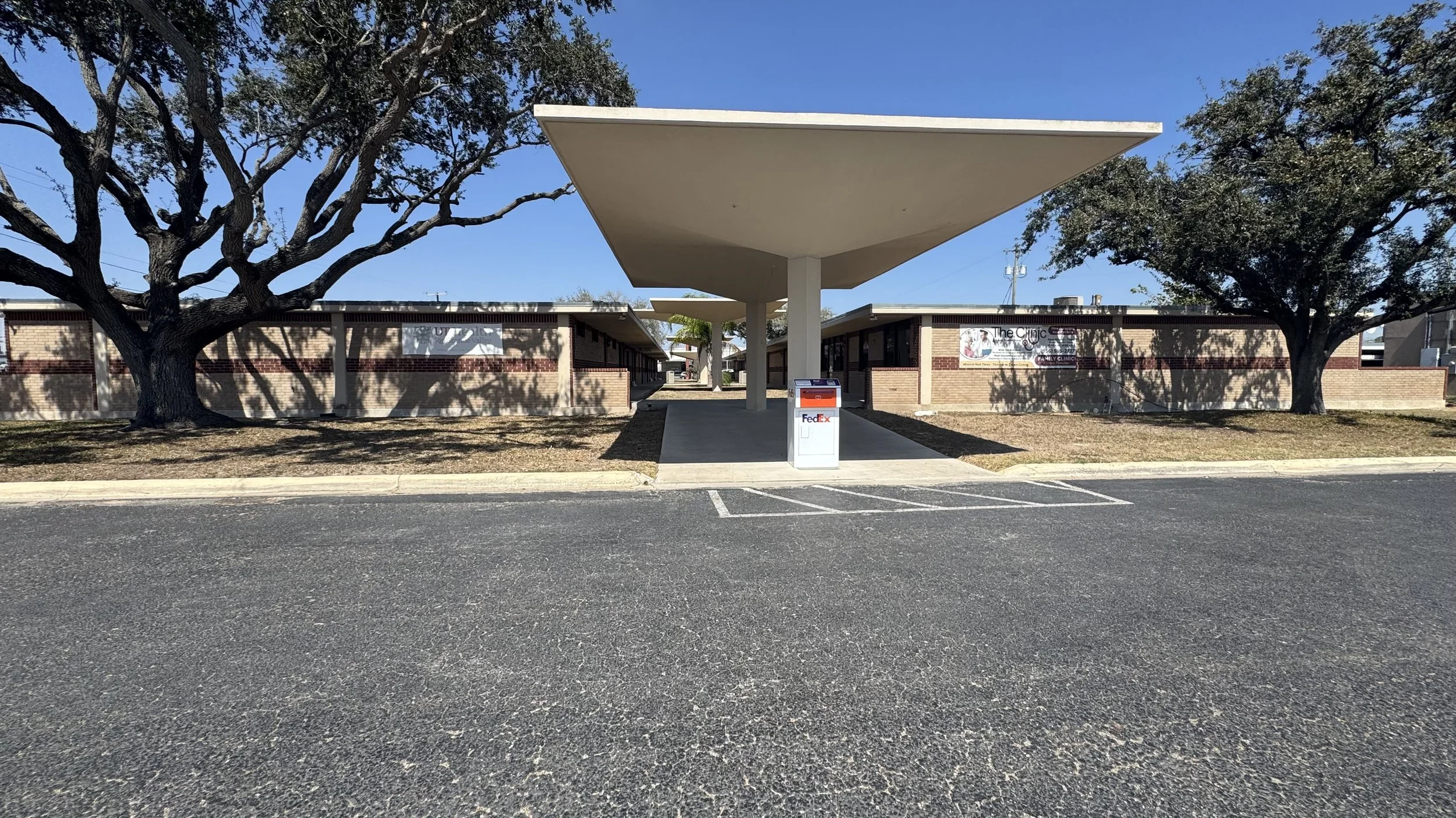 A single-story brick building with a large, modern white canopy in front, providing shade over walkways. There are two trees on either side of the building and a FedEx drop box in the middle of the sidewalk. The scene is outdoors on a sunny day with clear blue skies.
