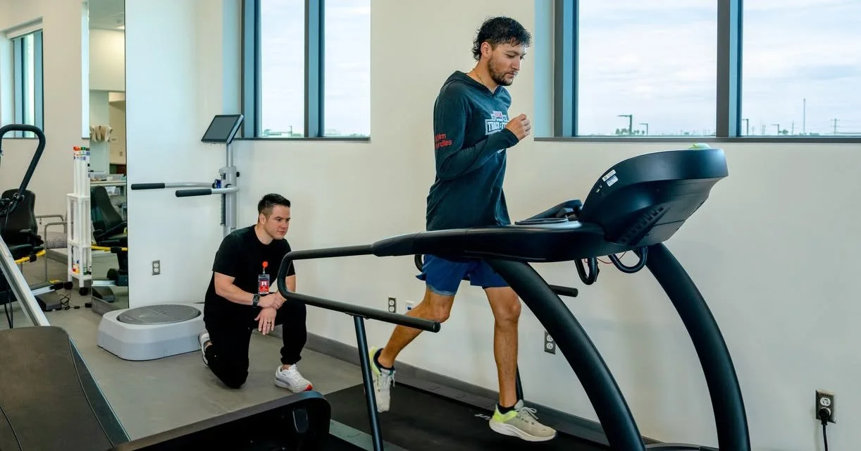 A man in athletic clothes running on a treadmill in a gym with two large windows in the background.