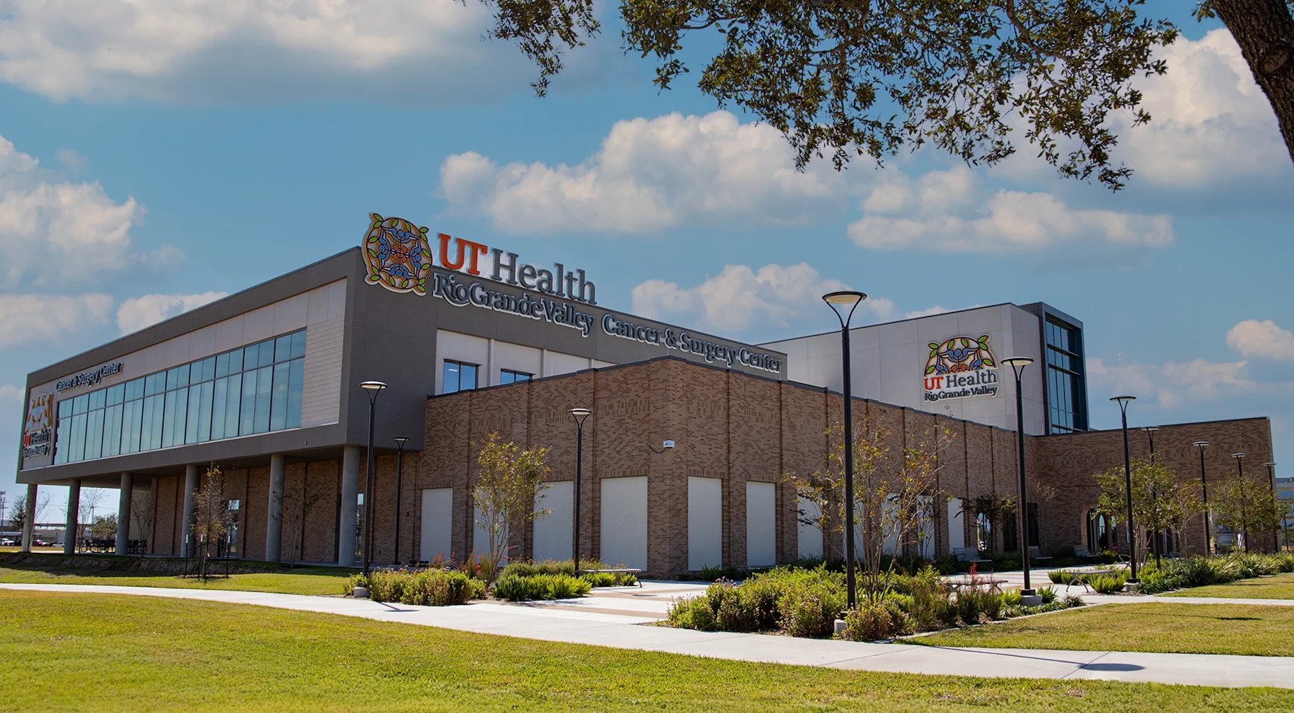 Exterior view of the UT Health Rio Grande Valley Cancer & Surgery Center building on a sunny day, with a blue sky and scattered clouds, surrounded by a landscaped lawn and small trees.