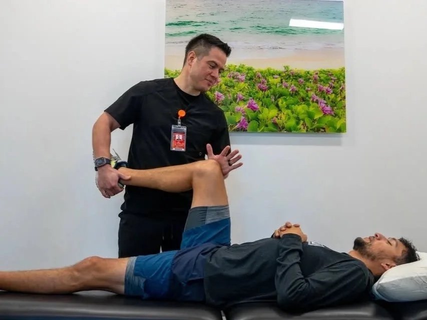 A physical therapist or trainer assisting a man with leg exercises on a therapy table in a medical or fitness setting, with a beach-themed picture on the wall.
