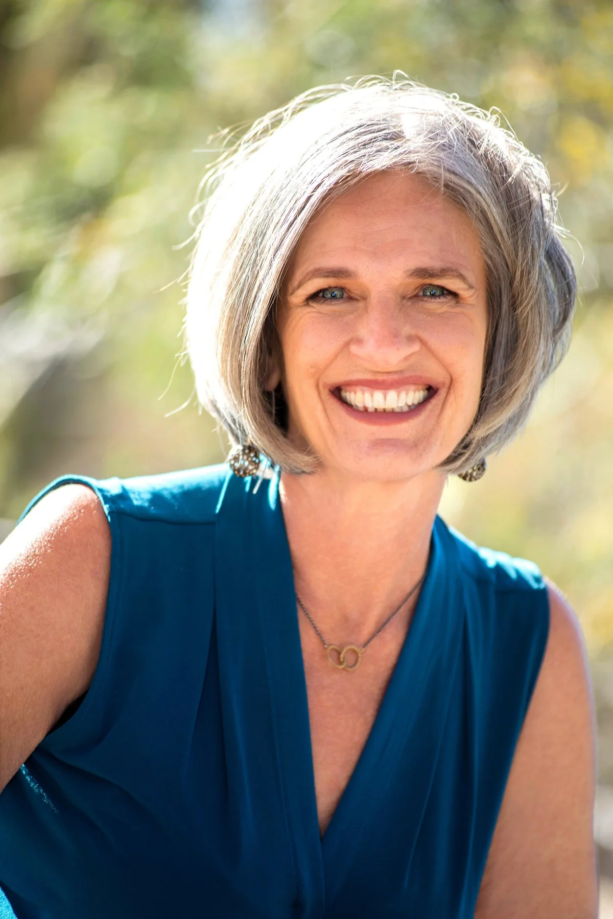 A smiling woman with short gray hair, wearing a blue sleeveless top, earrings, and a necklace, outdoors with a blurred natural background.