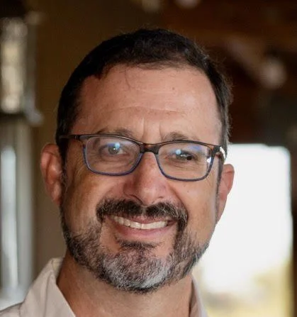 A middle-aged man with glasses, a beard, and mustache smiling indoors.