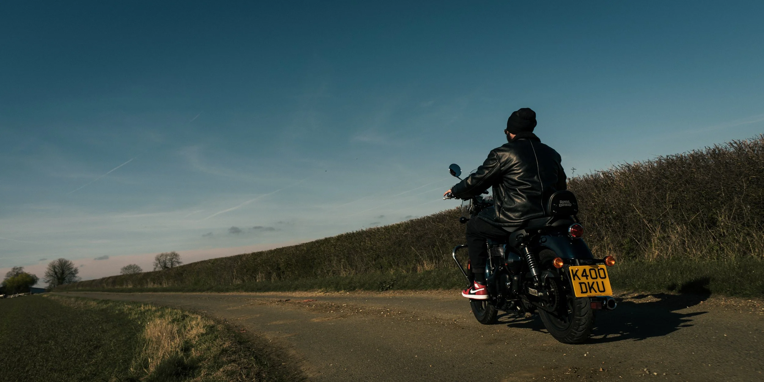 Person riding a black motorcycle on a rural road during sunset or sunrise, wearing a leather jacket and cap, with trees and open sky in the background.