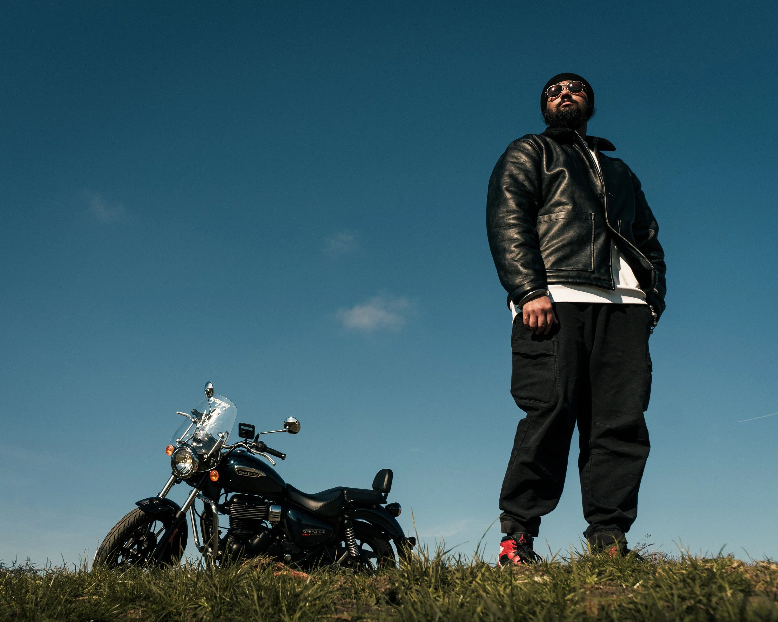 A man stands outdoors wearing a black leather jacket, black pants, sunglasses, and a beanie, next to a black motorcycle on a grassy field with a clear blue sky in the background.