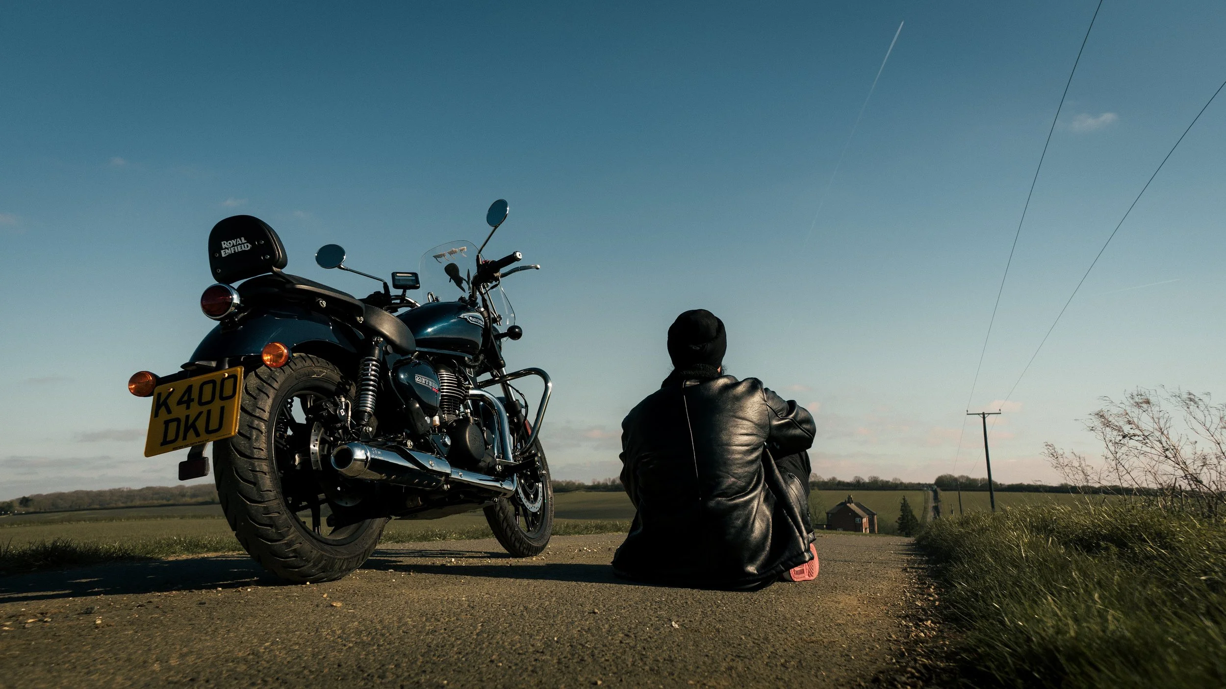 A person in a leather jacket sitting on a rural road next to a parked black motorcycle during sunset or sunrise.