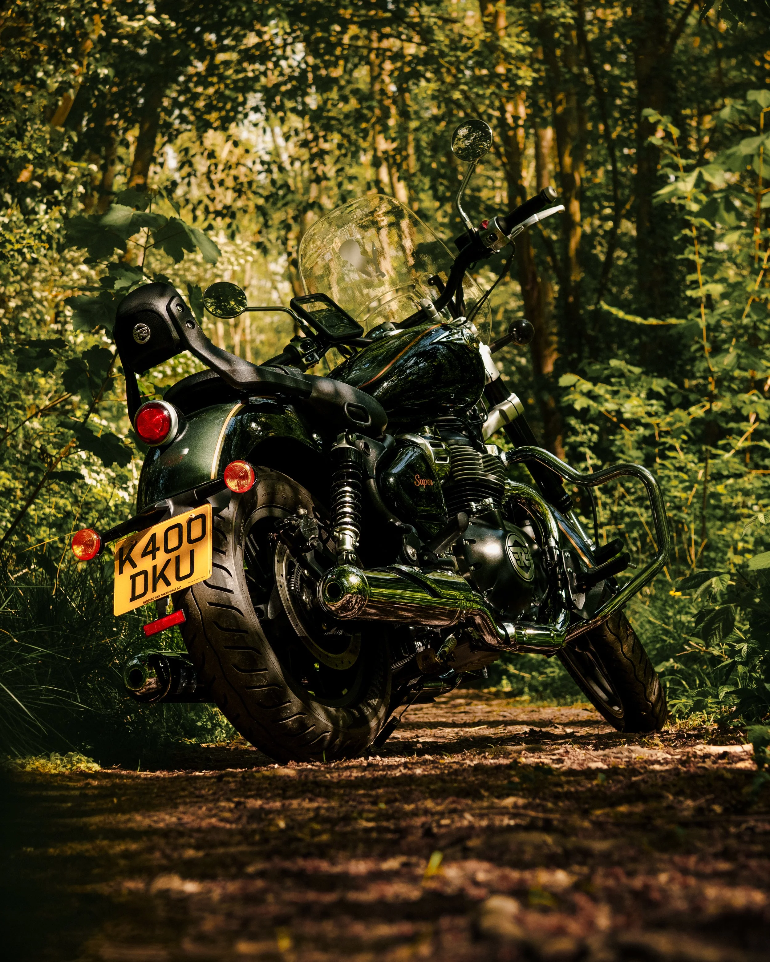 A black motorcycle with a yellow license plate that reads 'K400 DKU', parked on a dirt path in a dense forest with green foliage and trees.