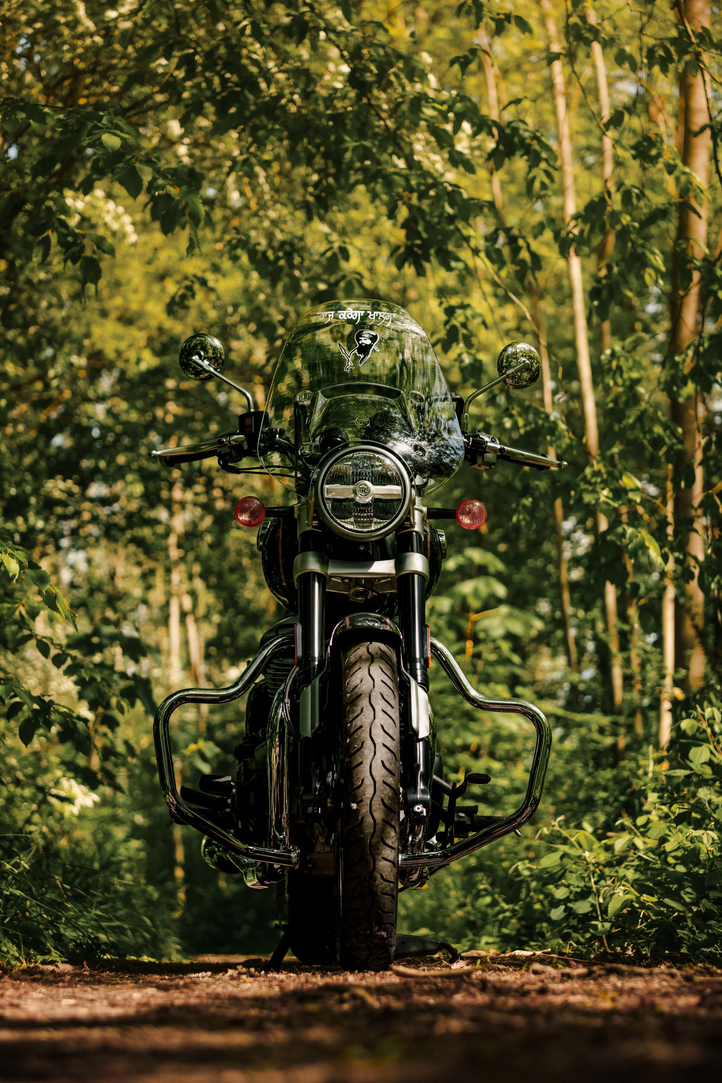 Front view of a black motorcycle parked on a dirt path surrounded by green trees and foliage in a forest setting.