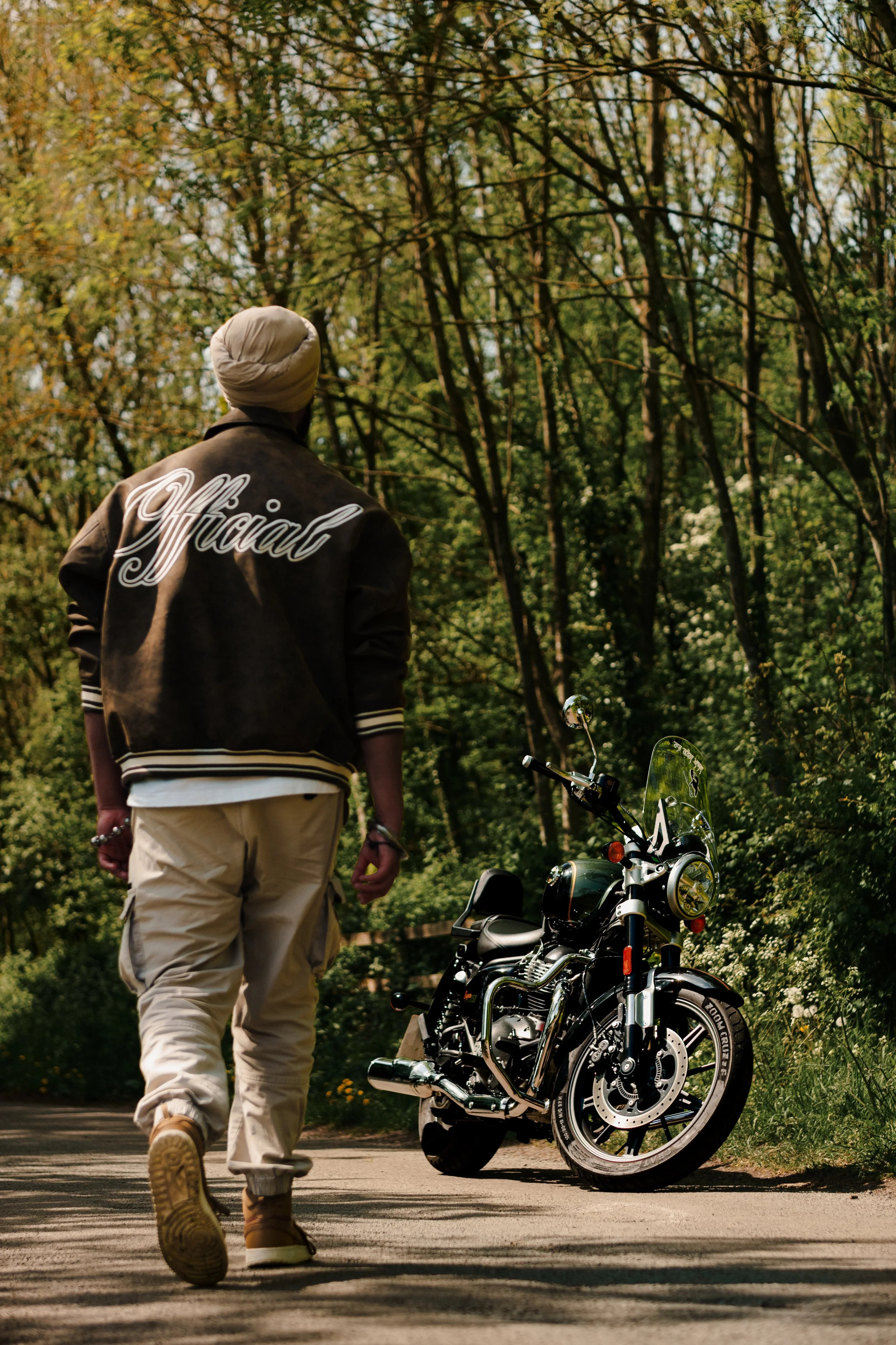 A person walking away from the camera on a forested path, wearing a jacket with 'Official' written on the back, with a motorcycle parked on the side of the path surrounded by tall trees and green foliage.