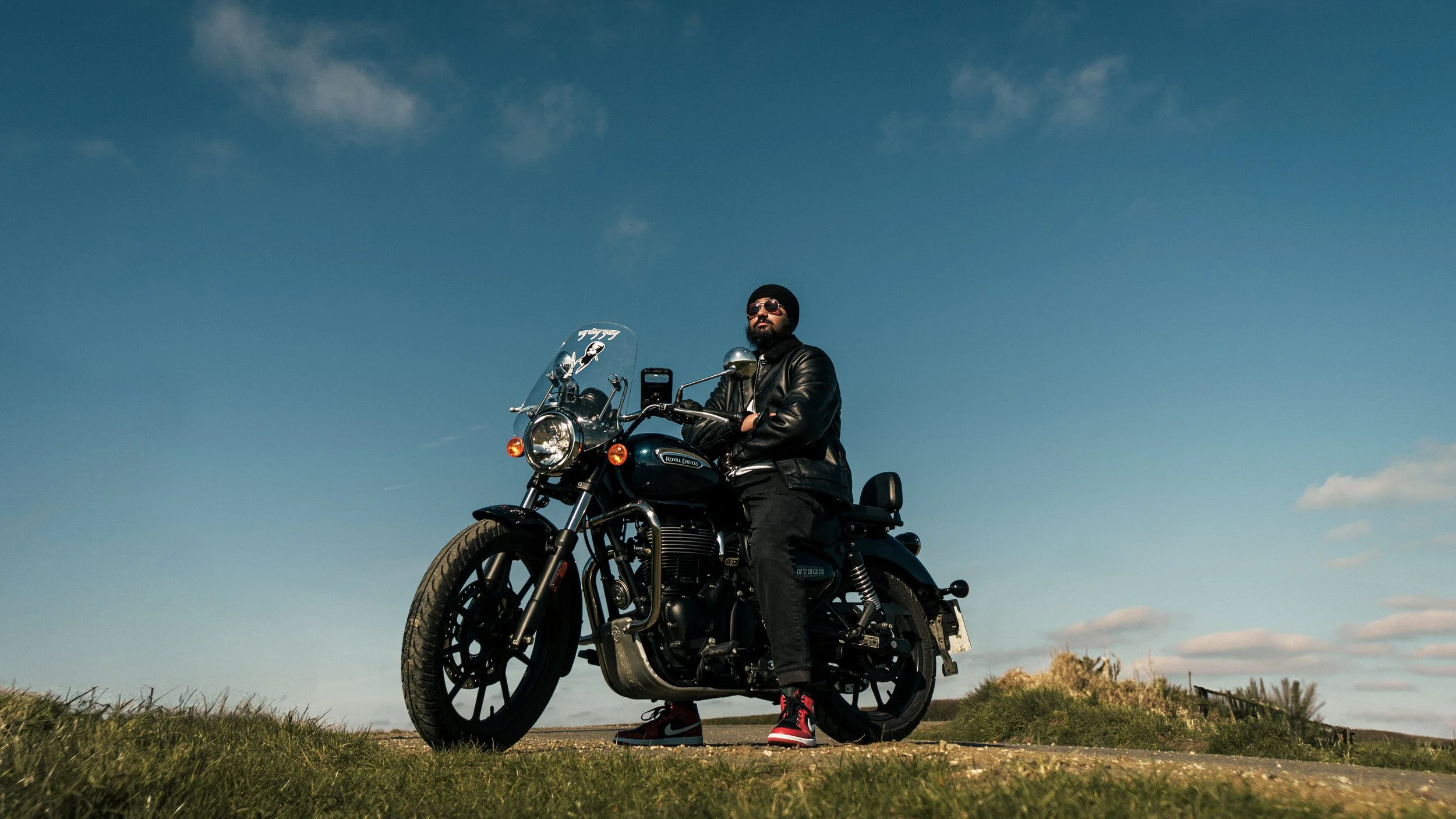 A man wearing sunglasses, a leather jacket, black pants, and red Nike sneakers sitting on a black motorcycle outdoors during daytime. The sky is clear with a few clouds and the man appears confident.