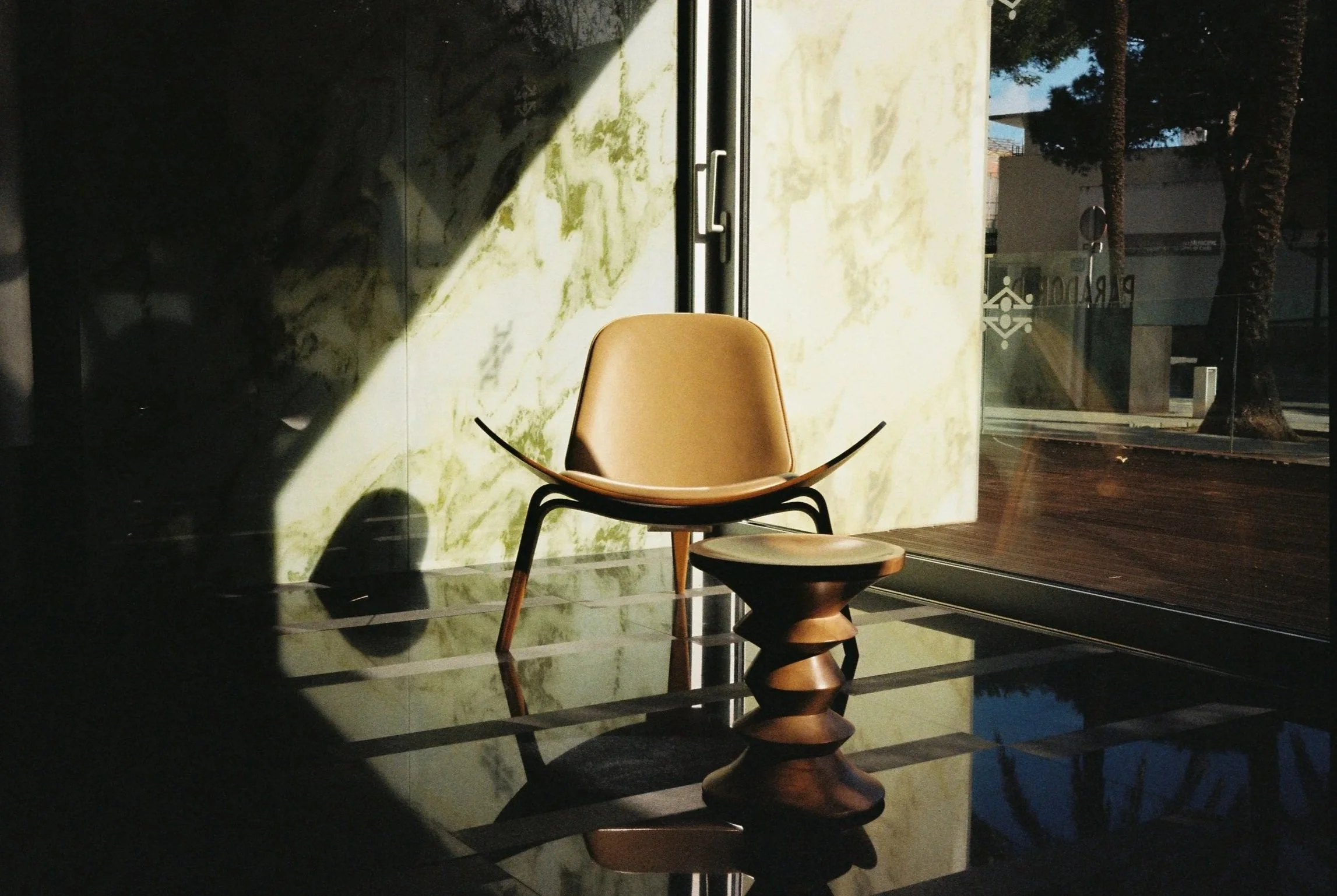 An interior scene with a single beige chair and a wooden side table placed on a reflective black tiled floor, illuminated by sunlight and cast shadows, with a window revealing trees and a building outside. Film photography by Anabela de Sousa