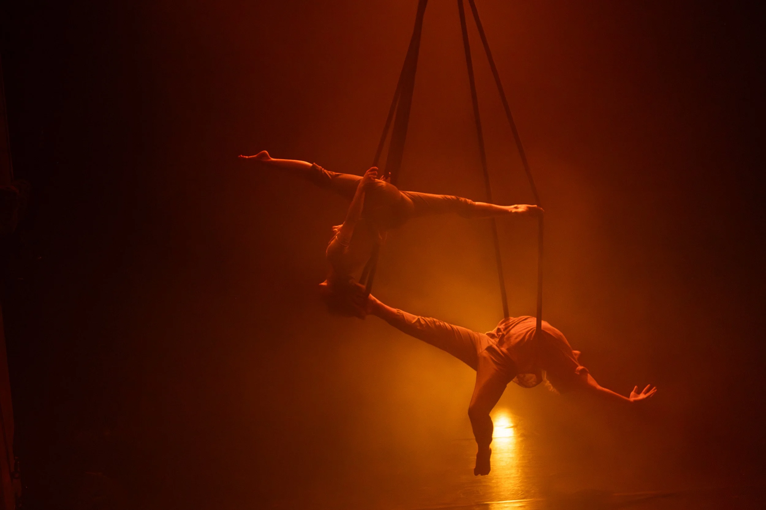 Two acrobats performing an aerial act on a trapeze with warm stage lighting.Performance Photography by Anabela de Sousa.