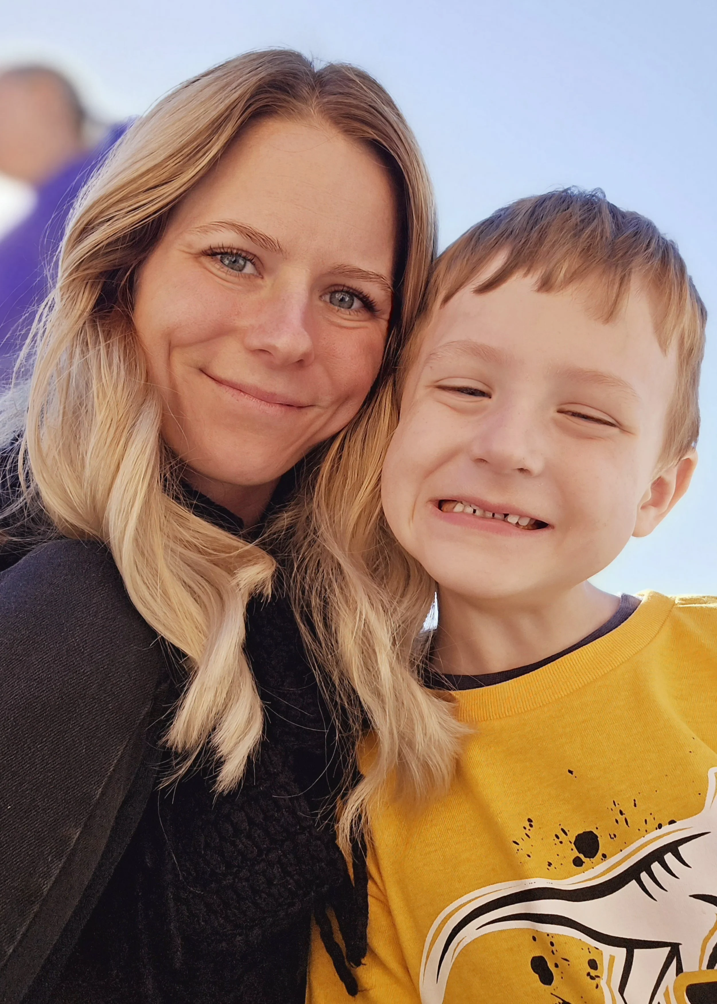 A woman and a boy smiling outdoors. The woman has blonde hair and blue eyes, and the boy has light brown hair and is wearing a yellow shirt with a graphic design.