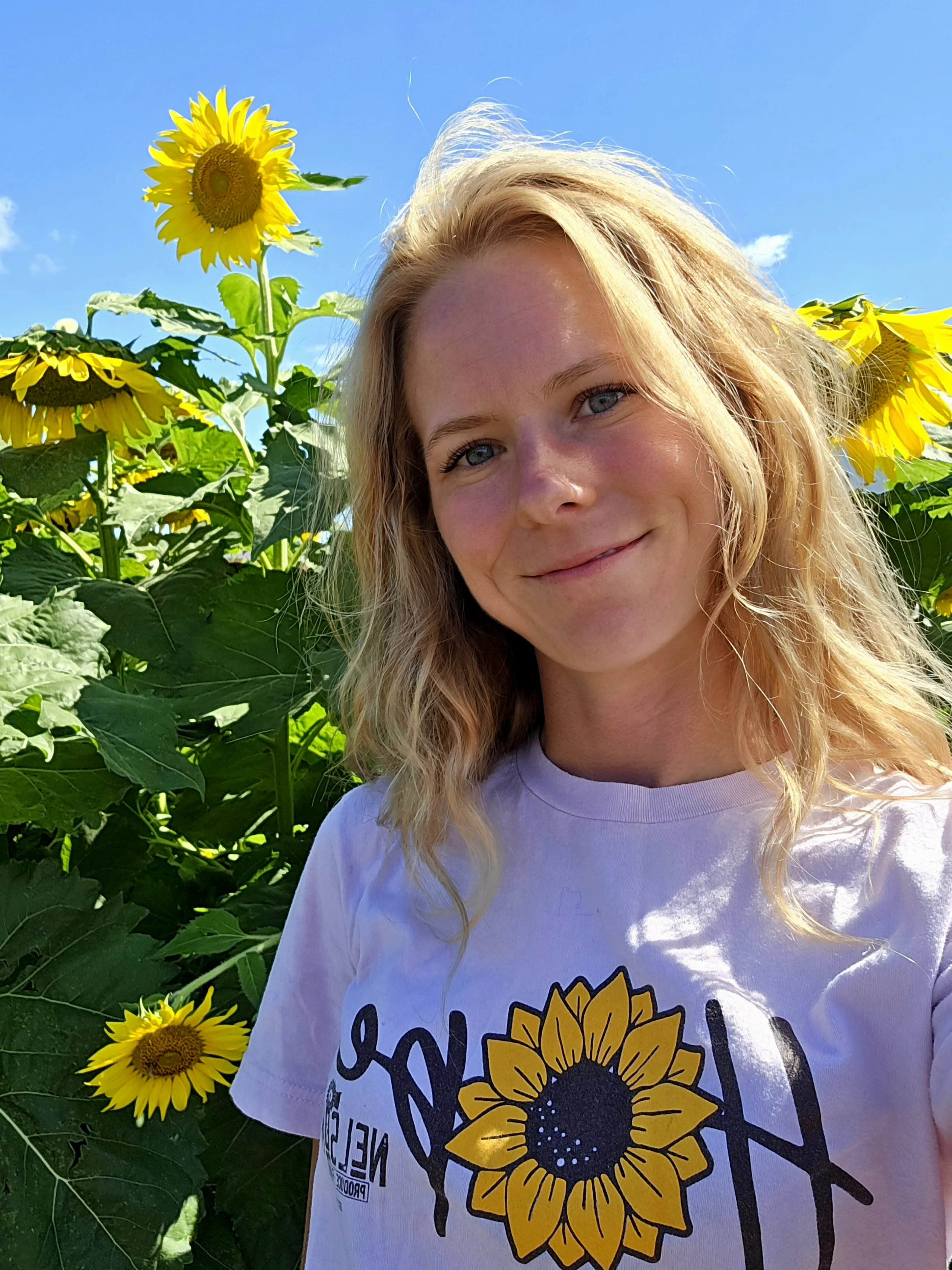 A young woman with wavy blonde hair smiling, standing in a sunflower field on a sunny day with blue sky and clouds in the background.