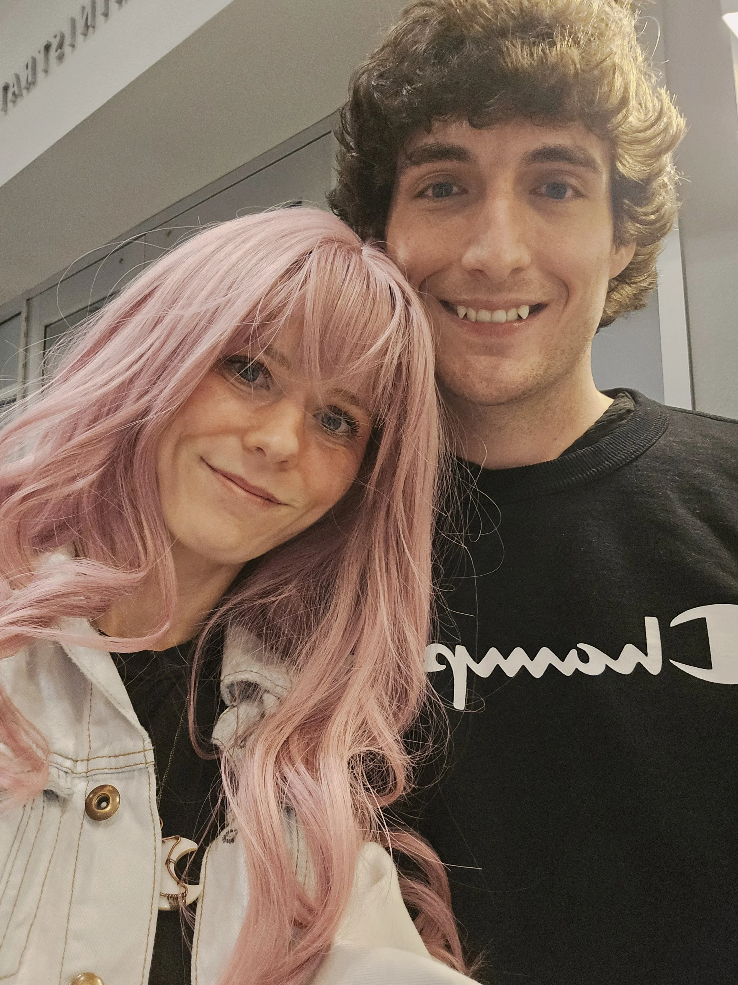 A woman with pink hair and a man with curly brown hair posing together for a selfie indoors.
