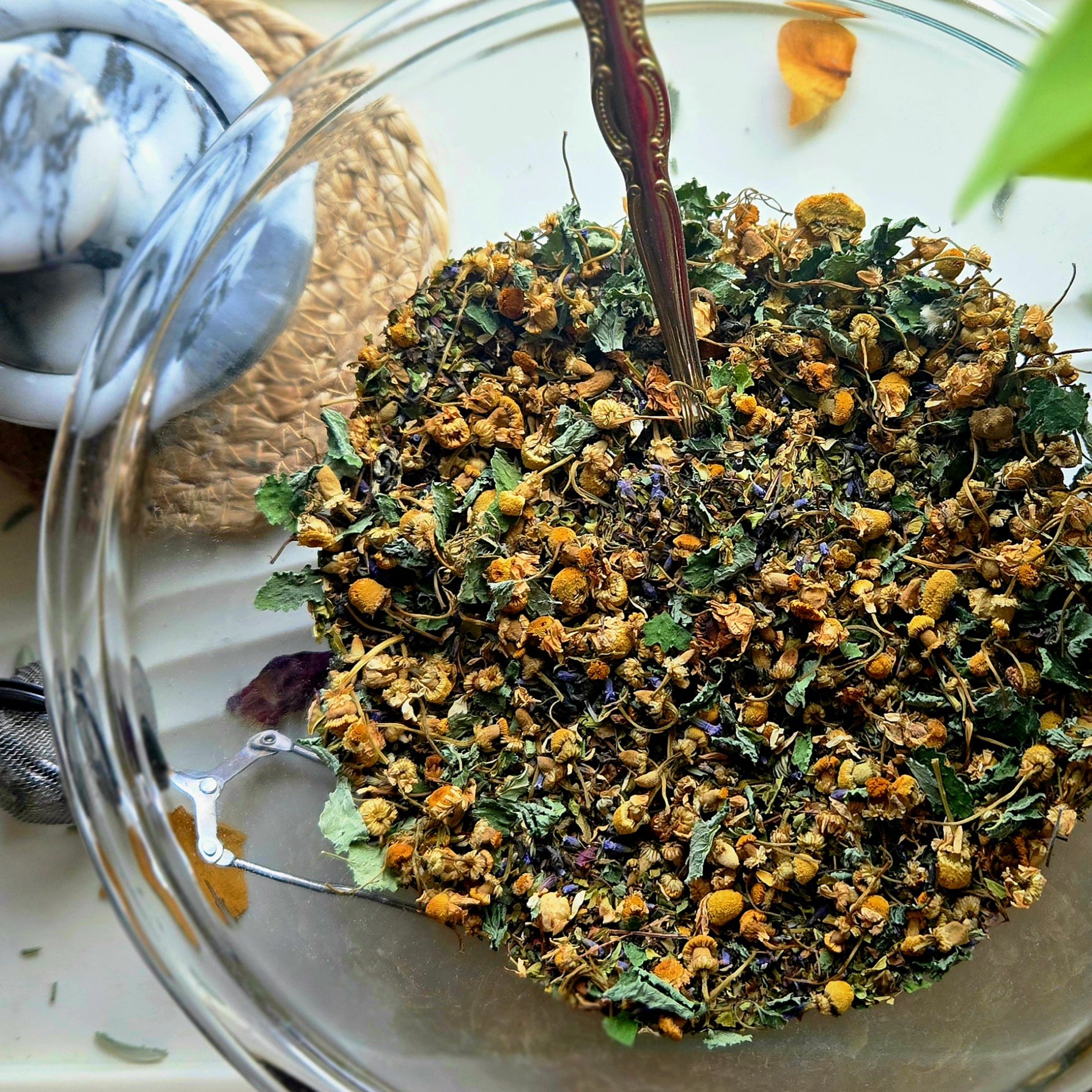 Dried chamomile flowers with leaves in a glass bowl, partially submerged in water, with a metal spoon resting among the flowers.