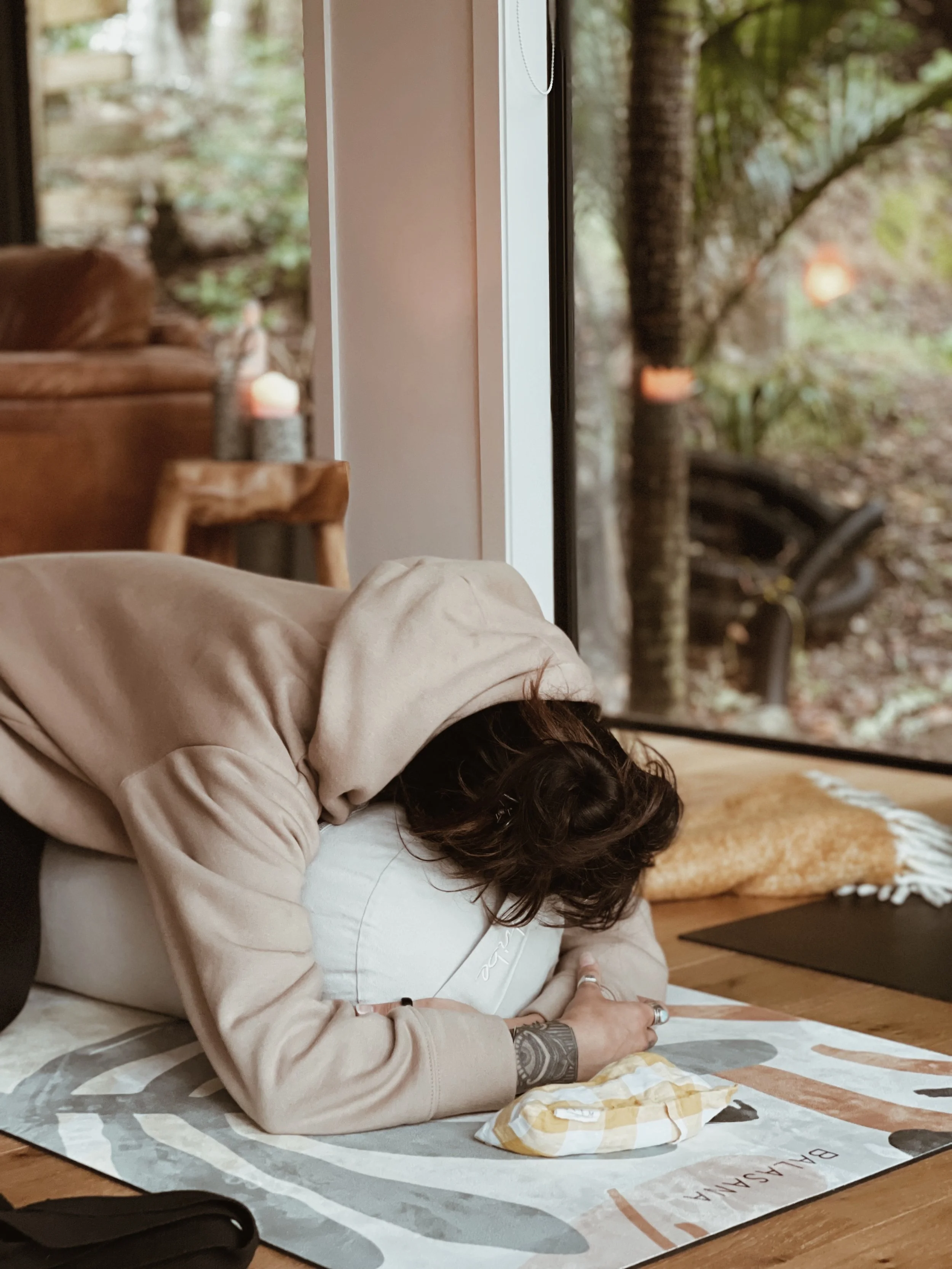 A person wearing a brown hoodie is lying on a yoga mat indoors, resting their head and arms on a cushion. The room has a cozy atmosphere with a view of greenery outside the window.
