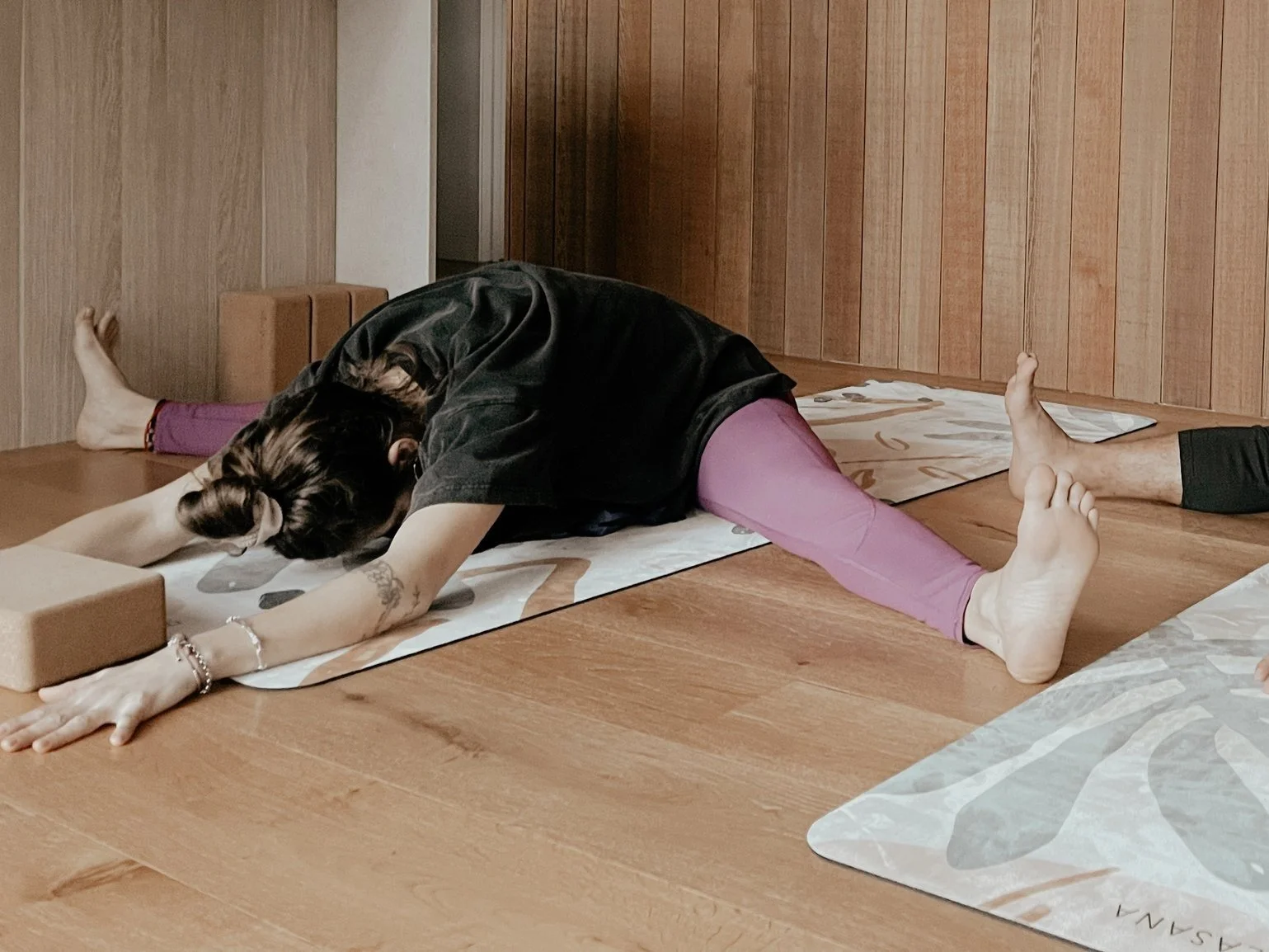Person doing a seated forward bend yoga pose on a mat, with arms stretched forward and legs spread wide, on a wooden floor.