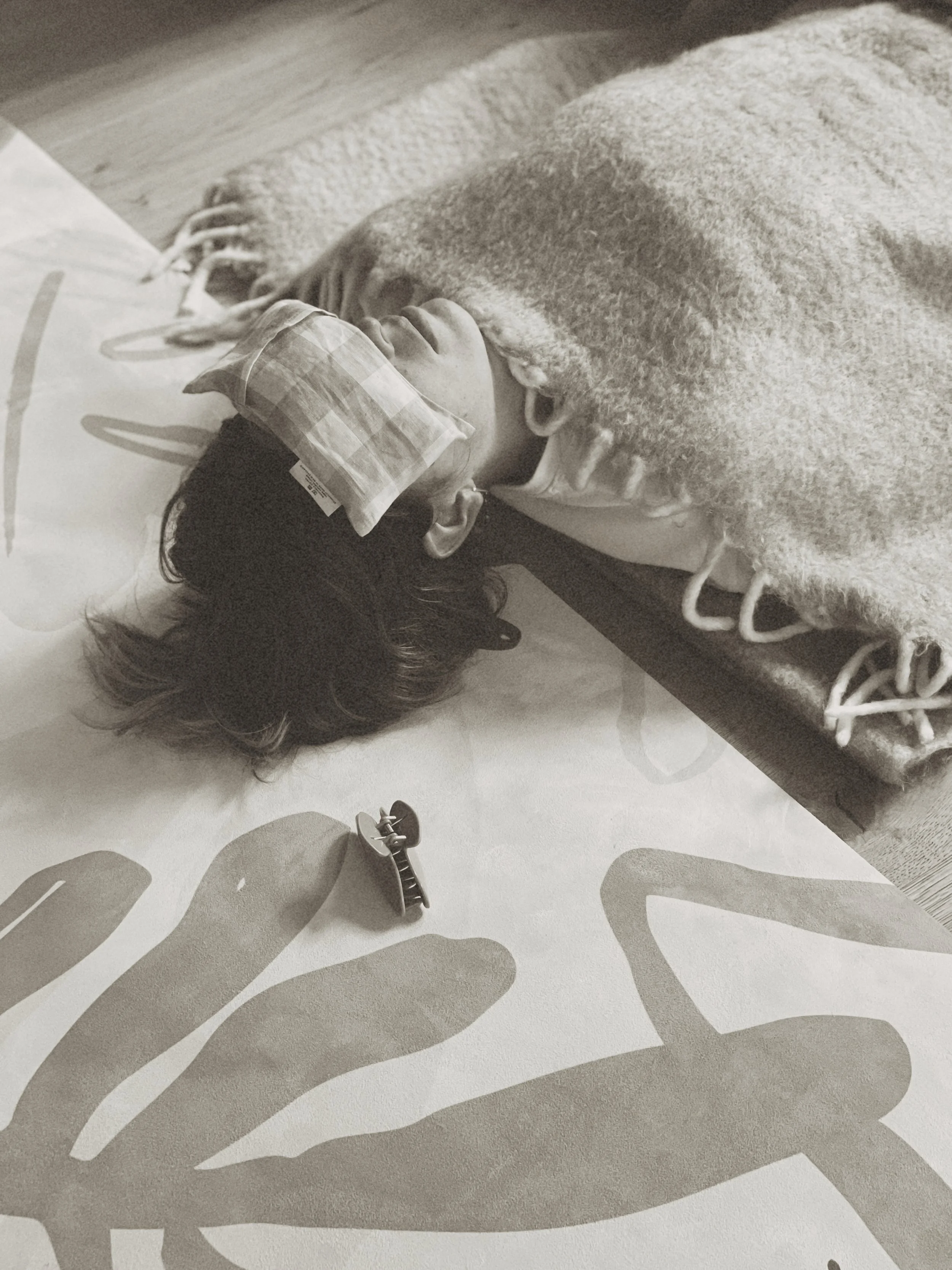 Person lying on a mat with an eye mask, covered by a blanket, in monochrome setting.